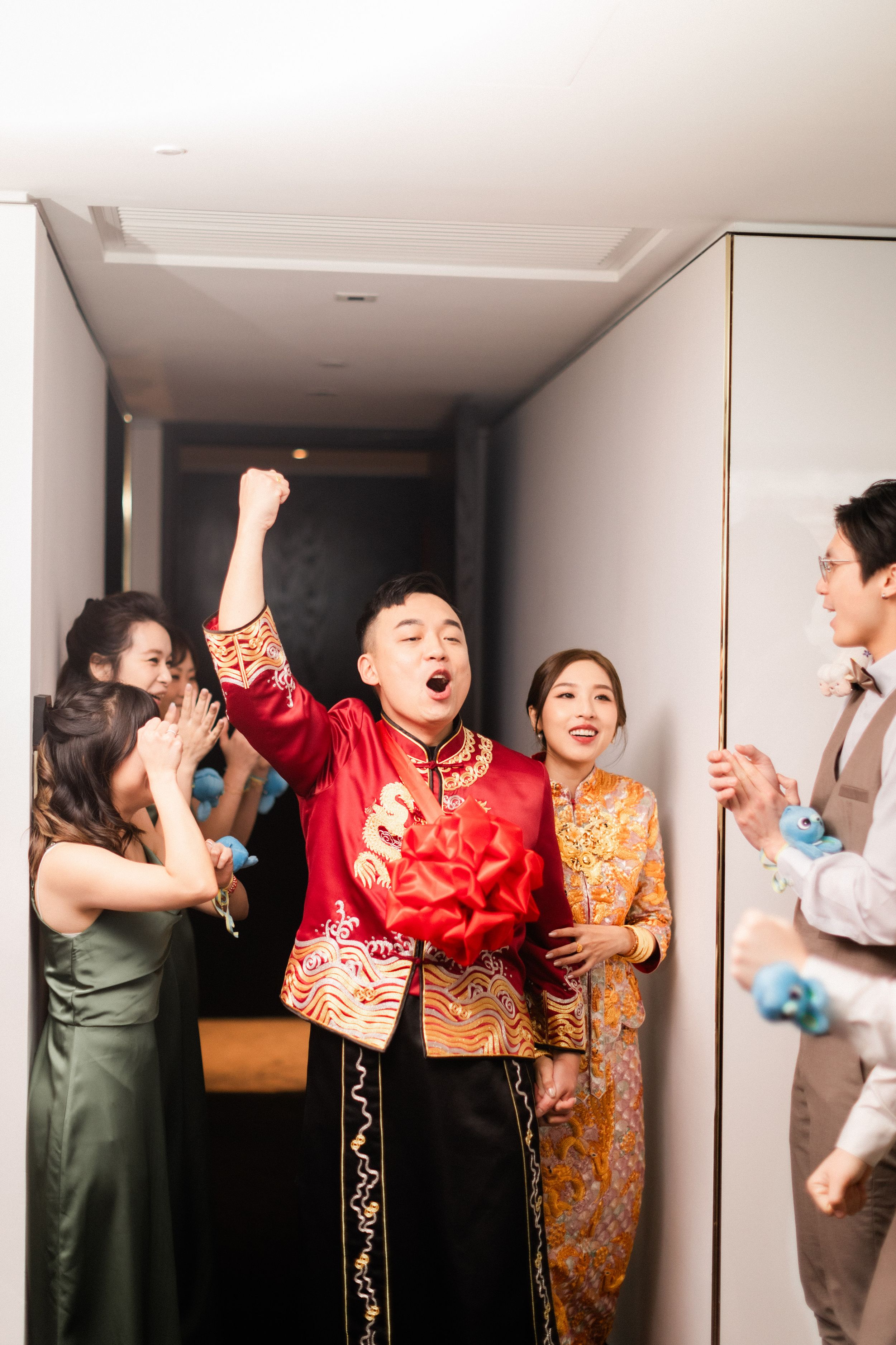 Excited groom in red Chinese wedding jacket celebrates with fist pump surrounded by wedding party in hotel corridor.