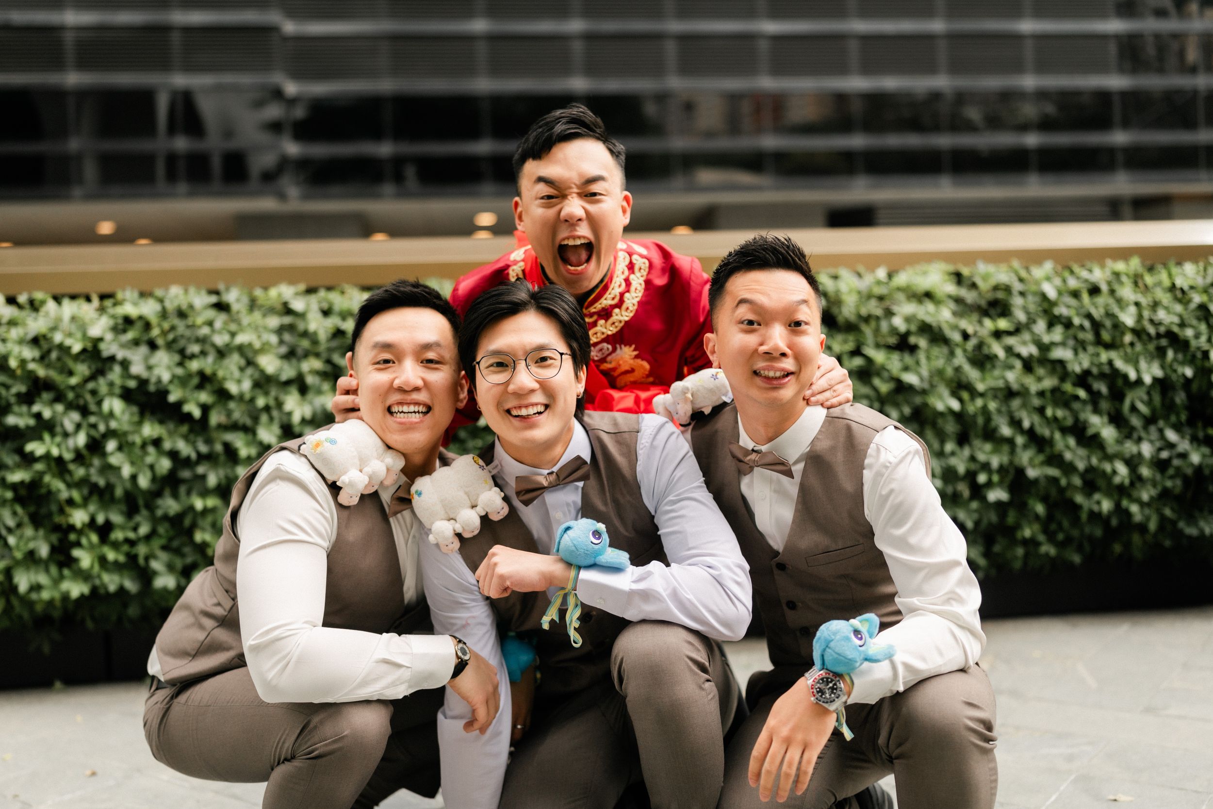 Groom with groomsmen in brown vests posing playfully outdoors in front of modern building.
