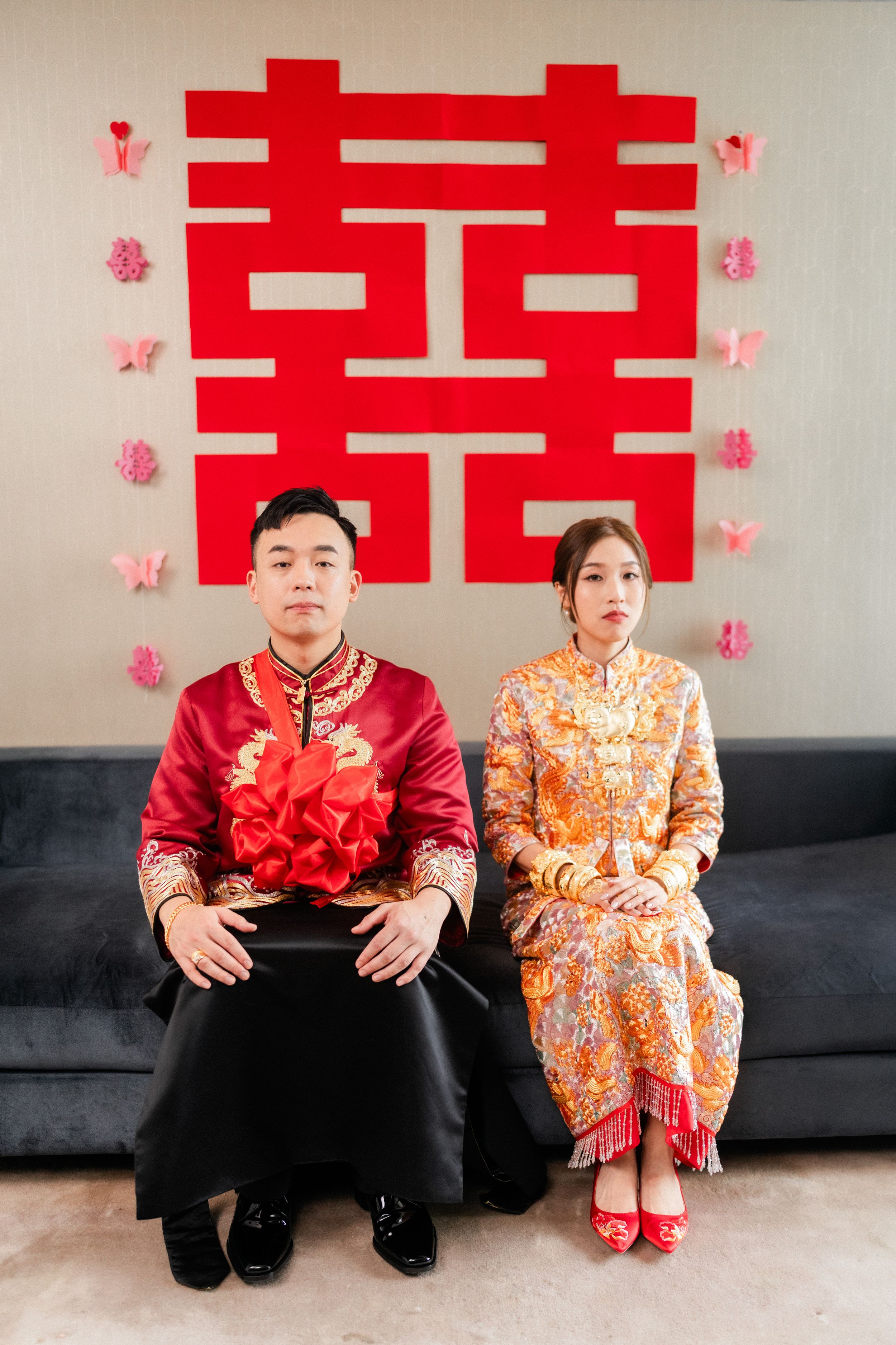 Chinese bride and groom in traditional red and gold wedding attire seated before large red double happiness symbol.