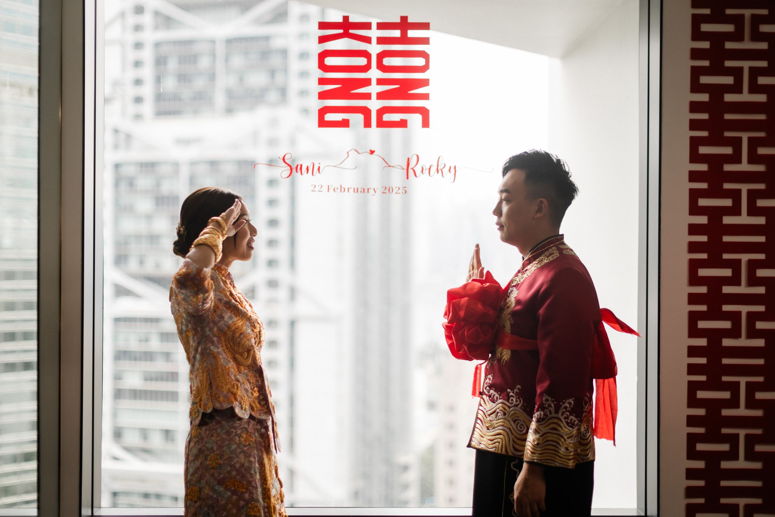Chinese bride and groom in traditional qipao attire exchange greetings by window with Hong Kong wedding signage.