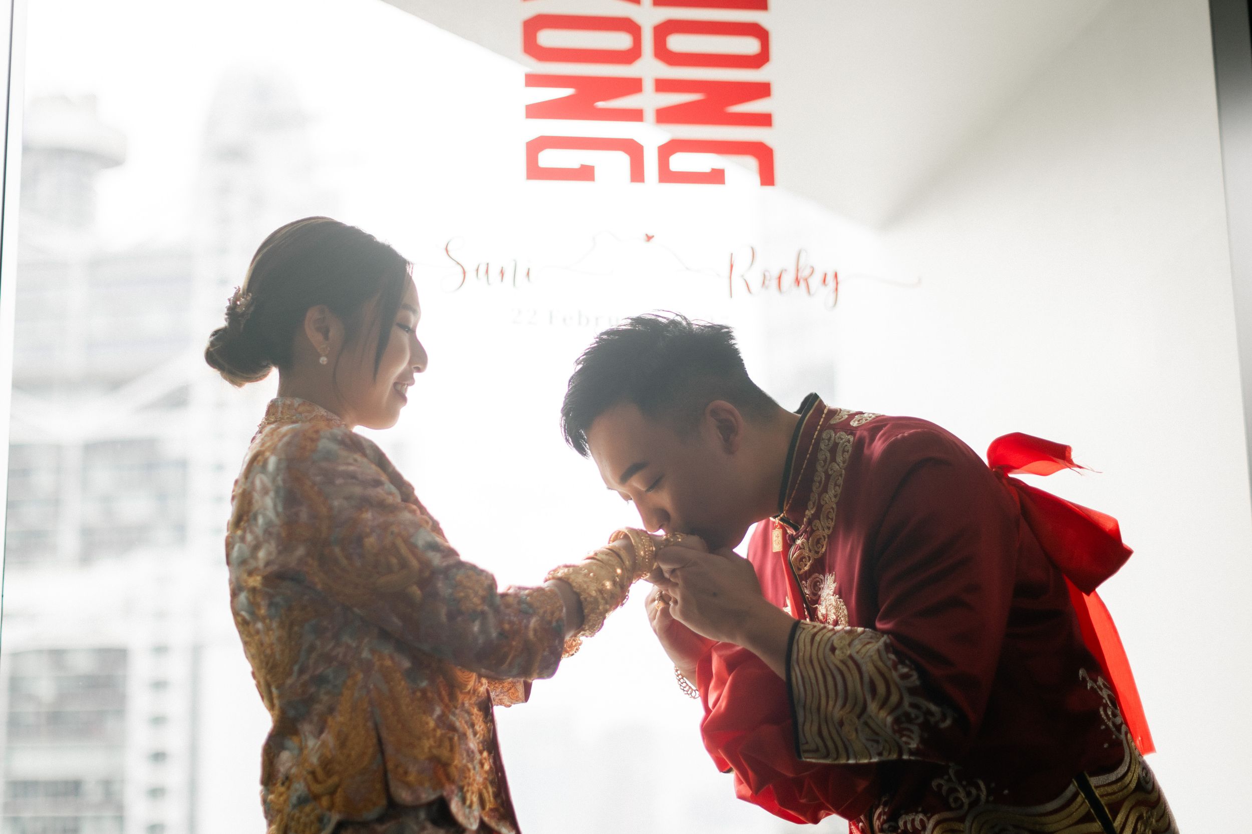 Chinese groom in red traditional wedding attire kisses bride's hand in tender romantic ceremony moment.