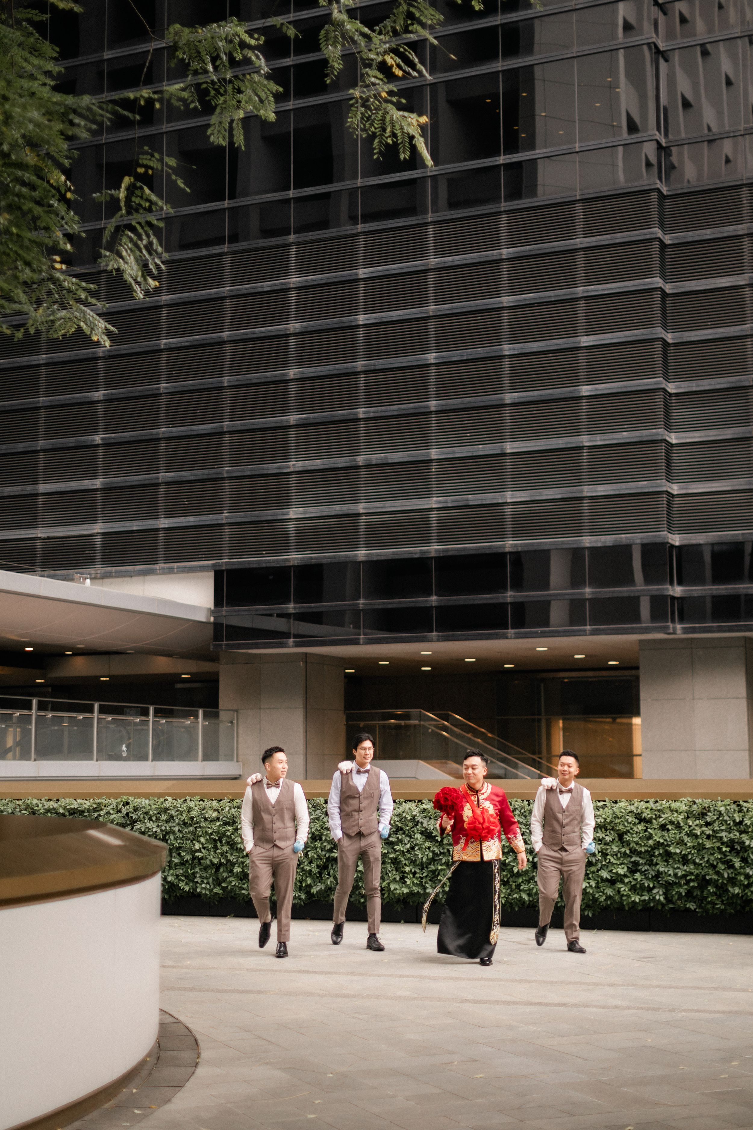 Bride in red qipao walking with groomsmen in brown vests outside modern glass building.