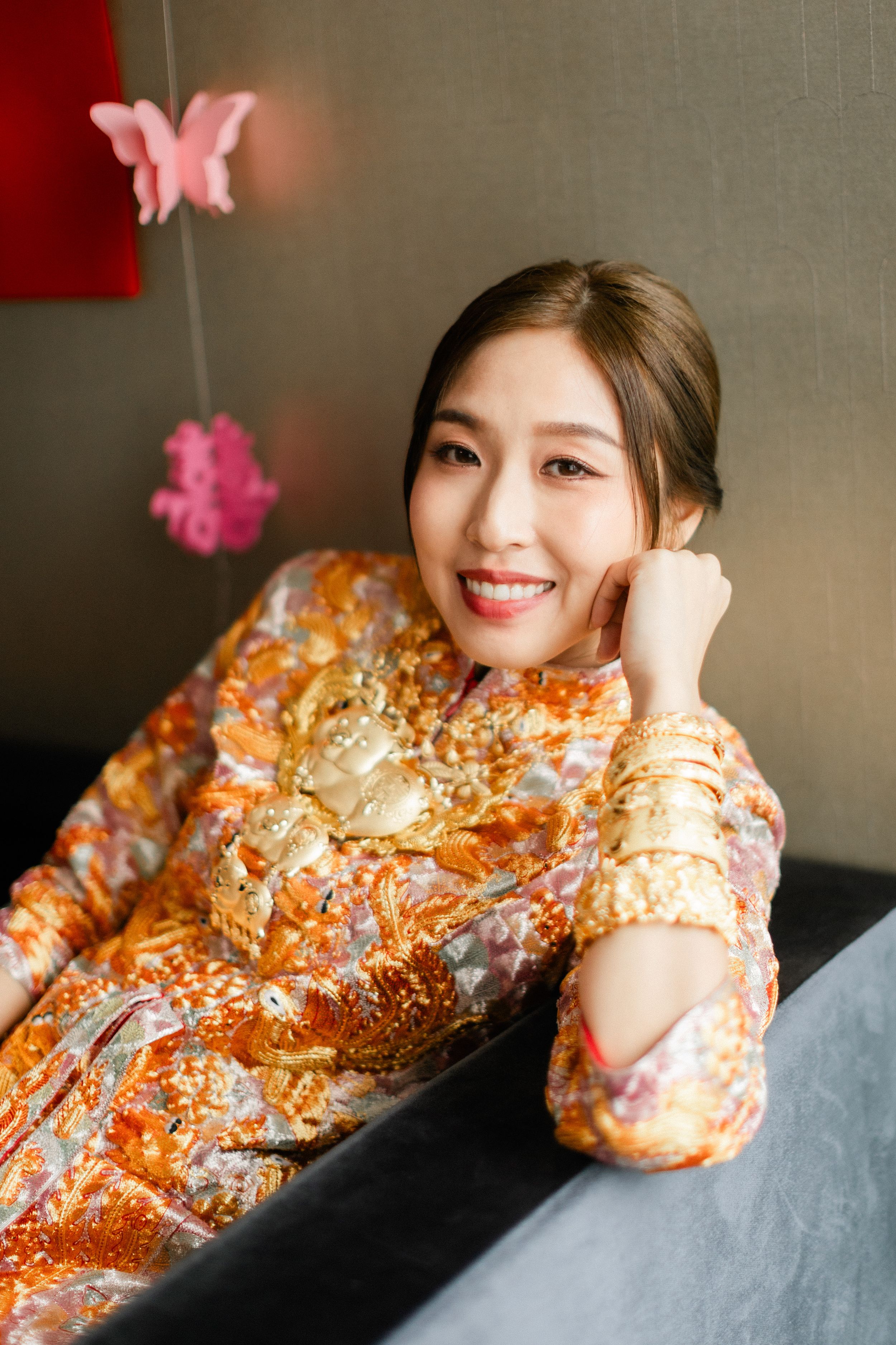 Beautiful Chinese bride in ornate gold and orange qipao adorned with traditional gold jewelry smiling elegantly.