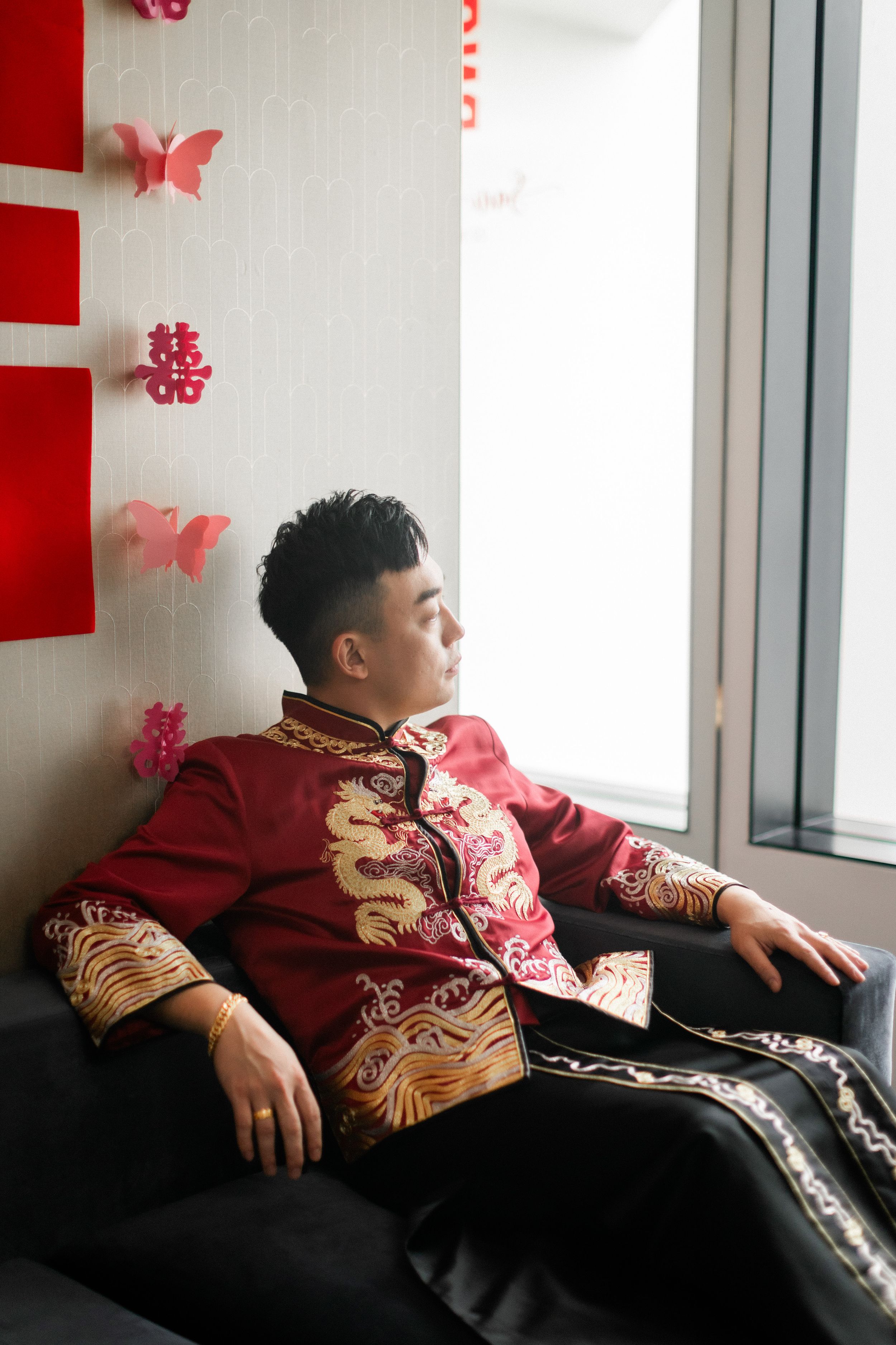 Chinese groom in red dragon embroidered traditional wedding jacket sits pensively by window with pink butterfly decor.