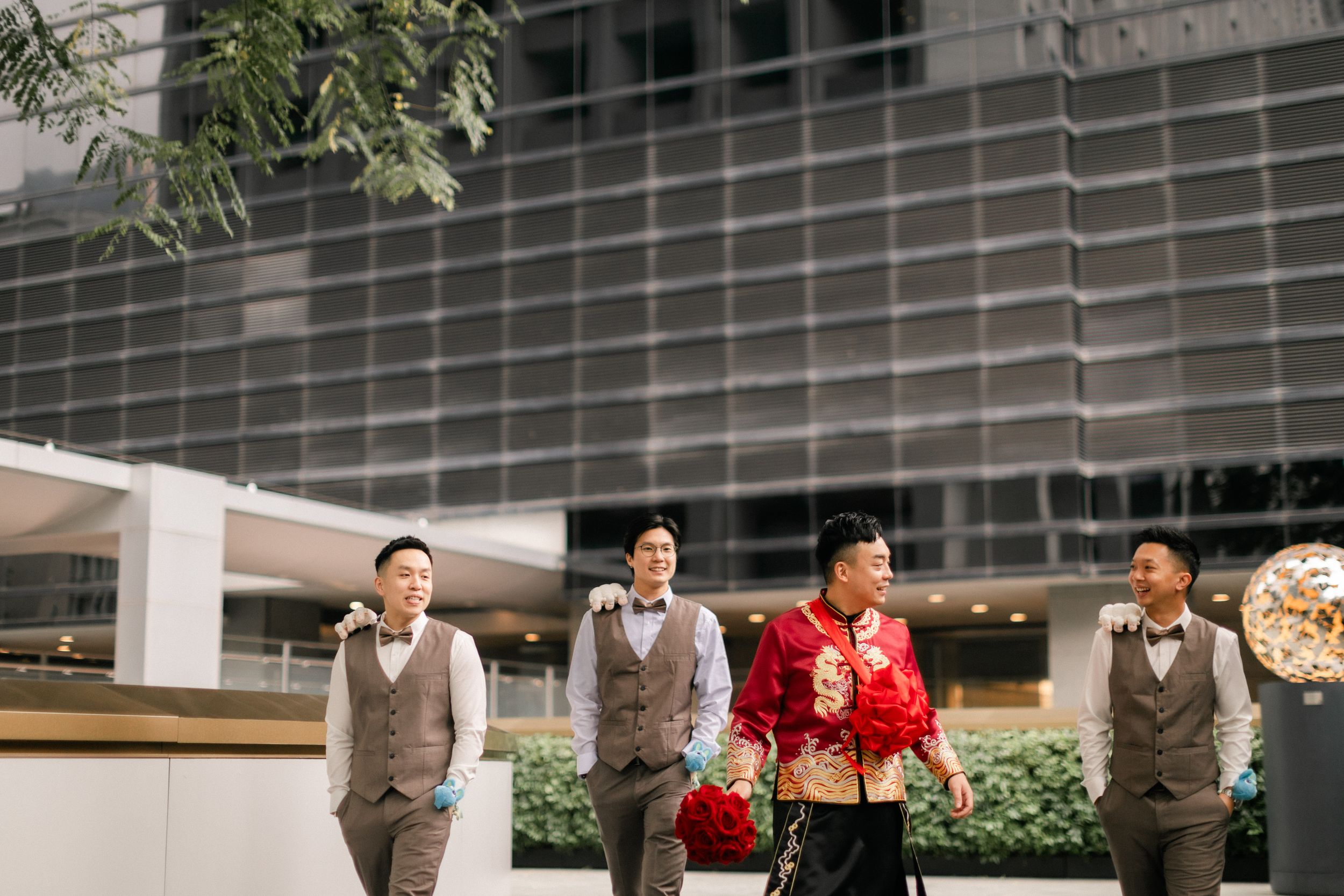 Groom in traditional red Chinese wedding attire walking with groomsmen outside modern building.