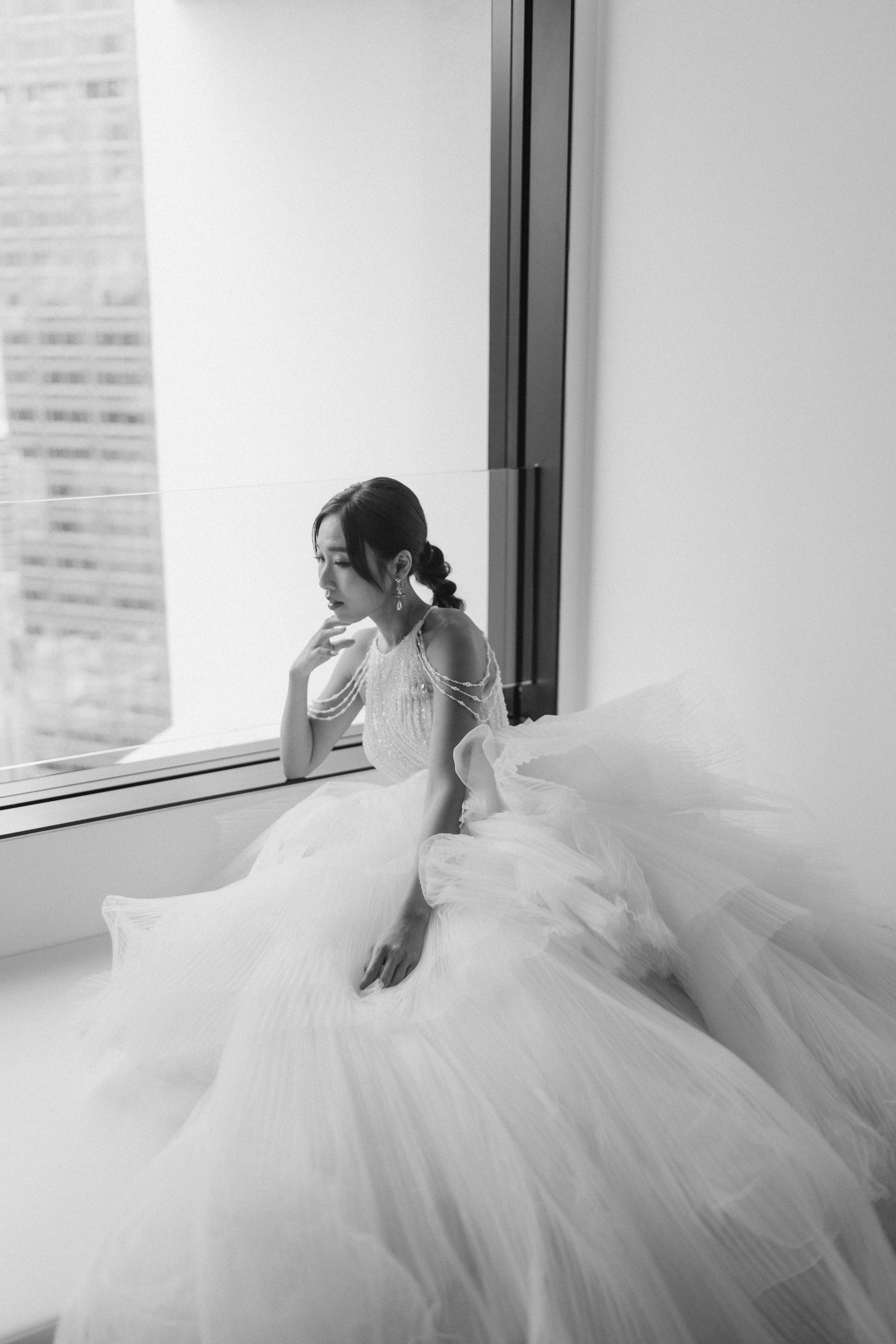 Elegant bride in a white tulle ball gown sitting by a large window in black and white photo.