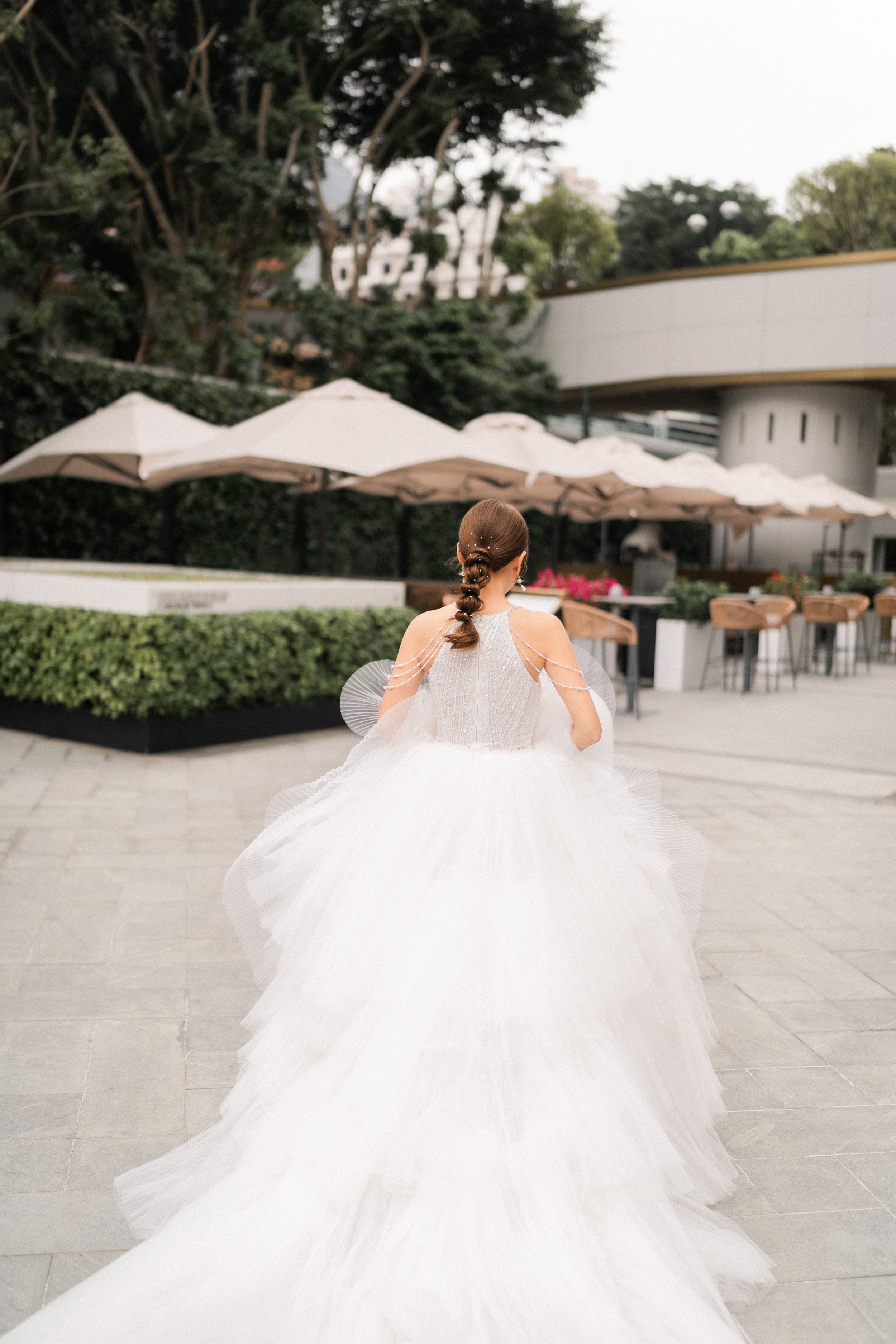 Bride in a layered white tulle wedding gown walking outdoors near a garden patio area from behind.