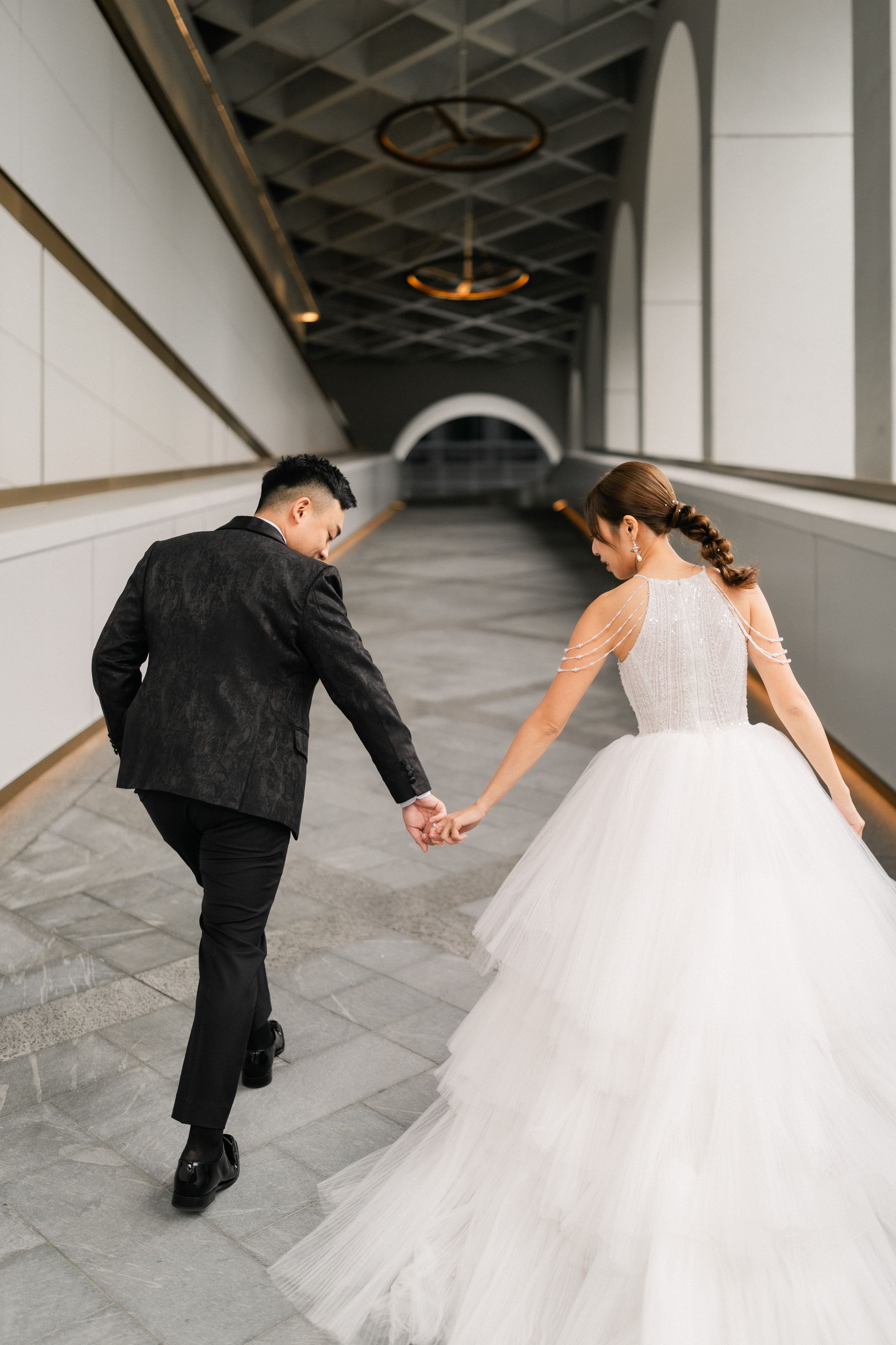 Bride and groom holding hands walking up steps in a modern architectural corridor from behind.