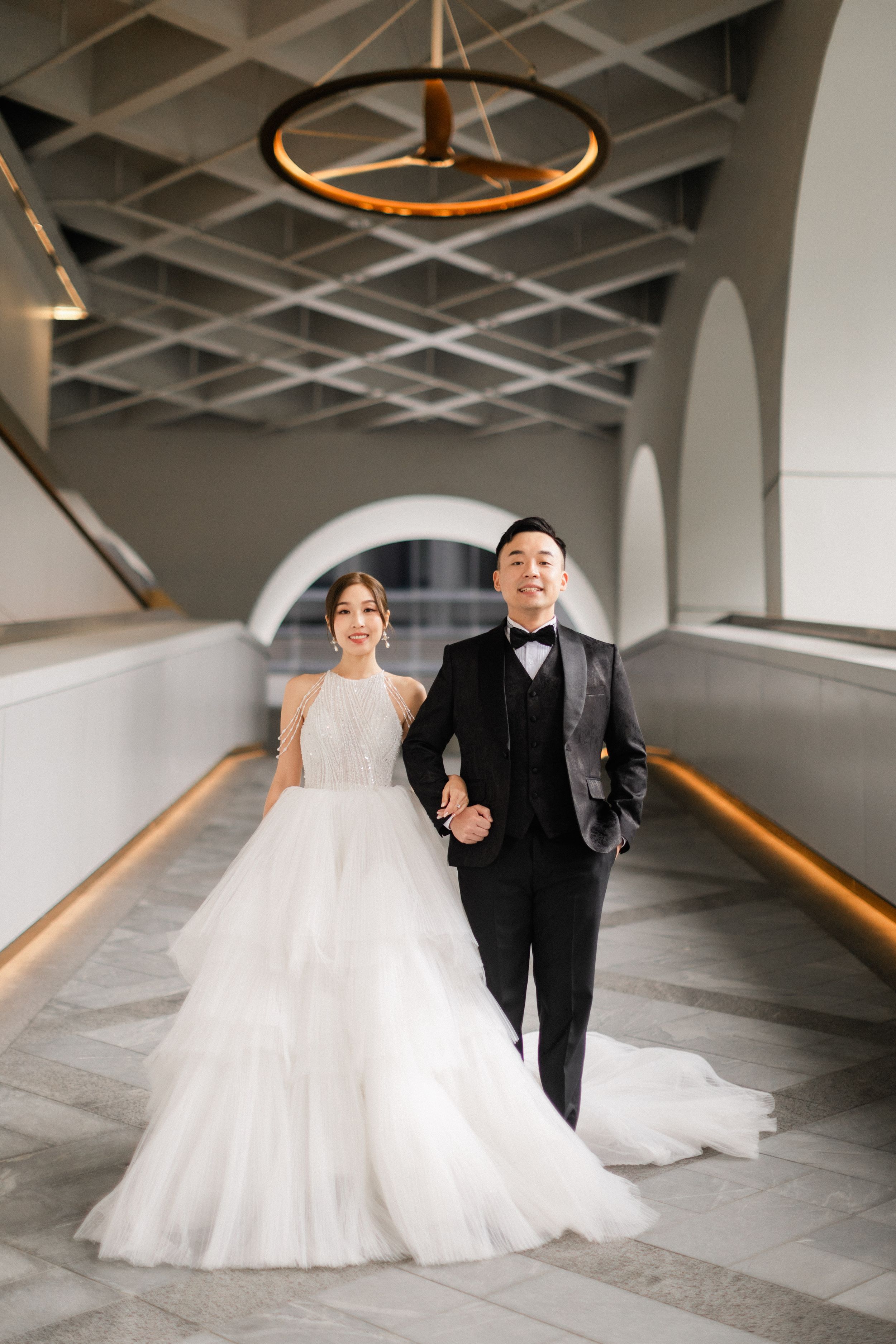 Asian couple in wedding attire posing in an elegant architectural hallway with arched ceiling details.