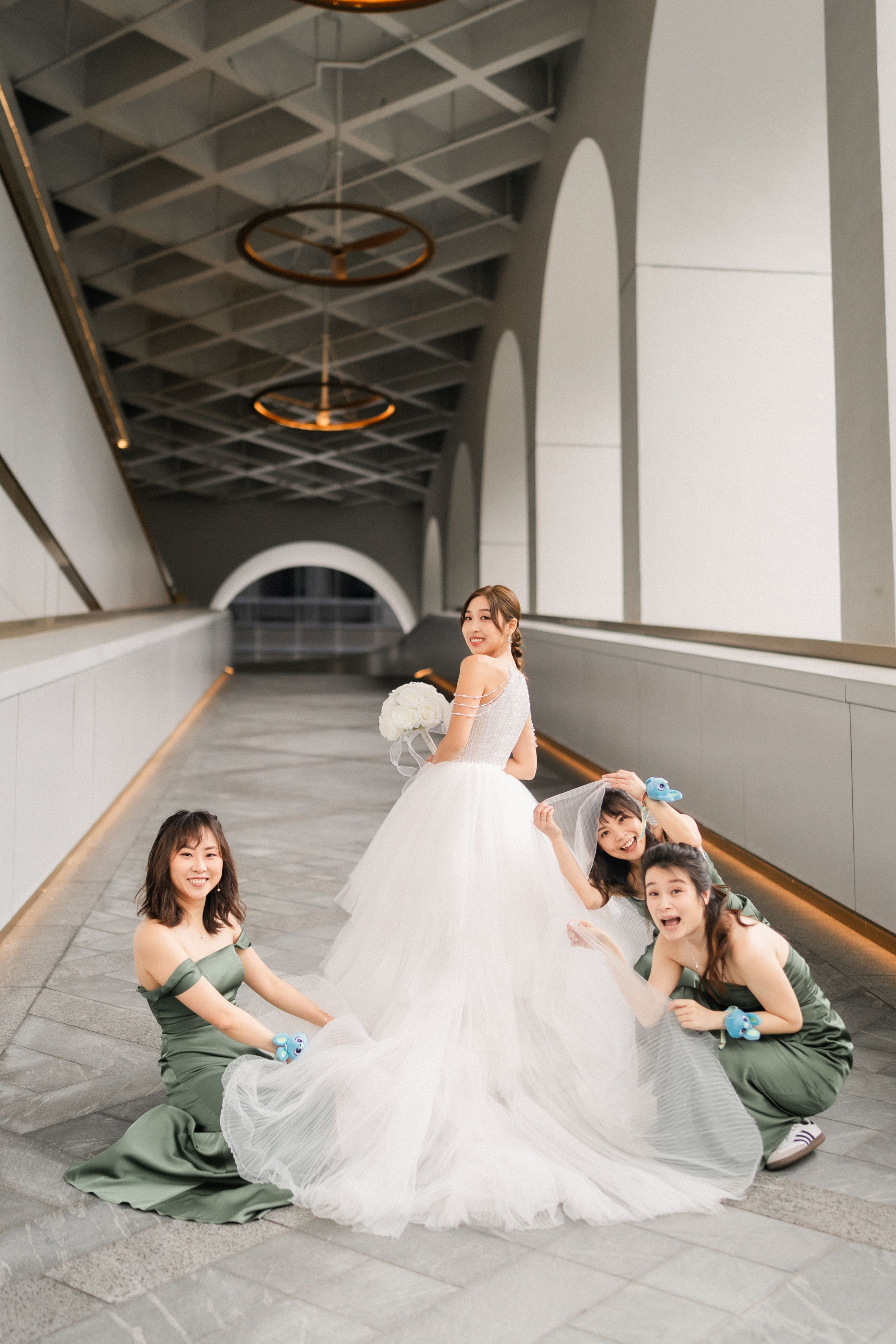 Bride in white ball gown posing with three bridesmaids in sage green dresses in an elegant corridor.