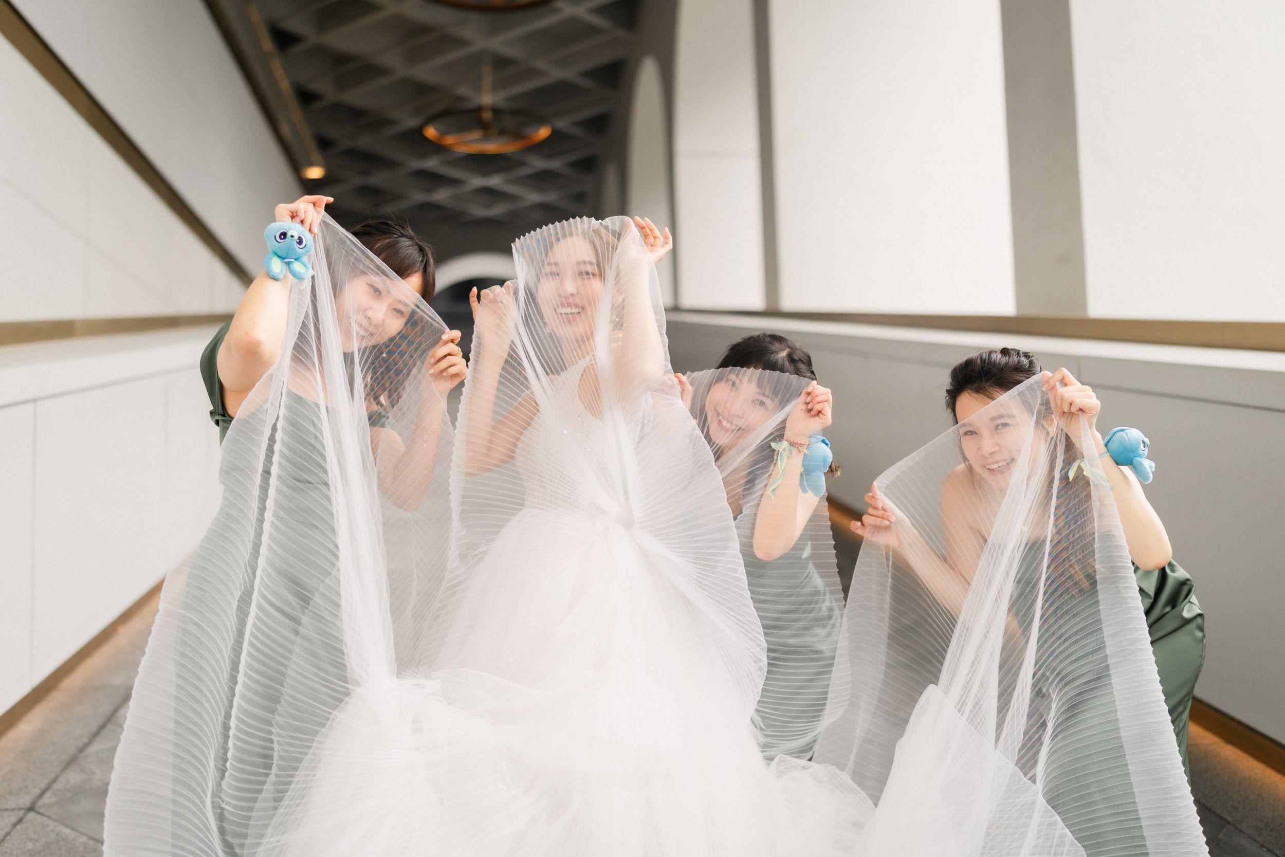 Bridesmaids in sage green dresses holding up the veil of a smiling bride in a bright corridor.