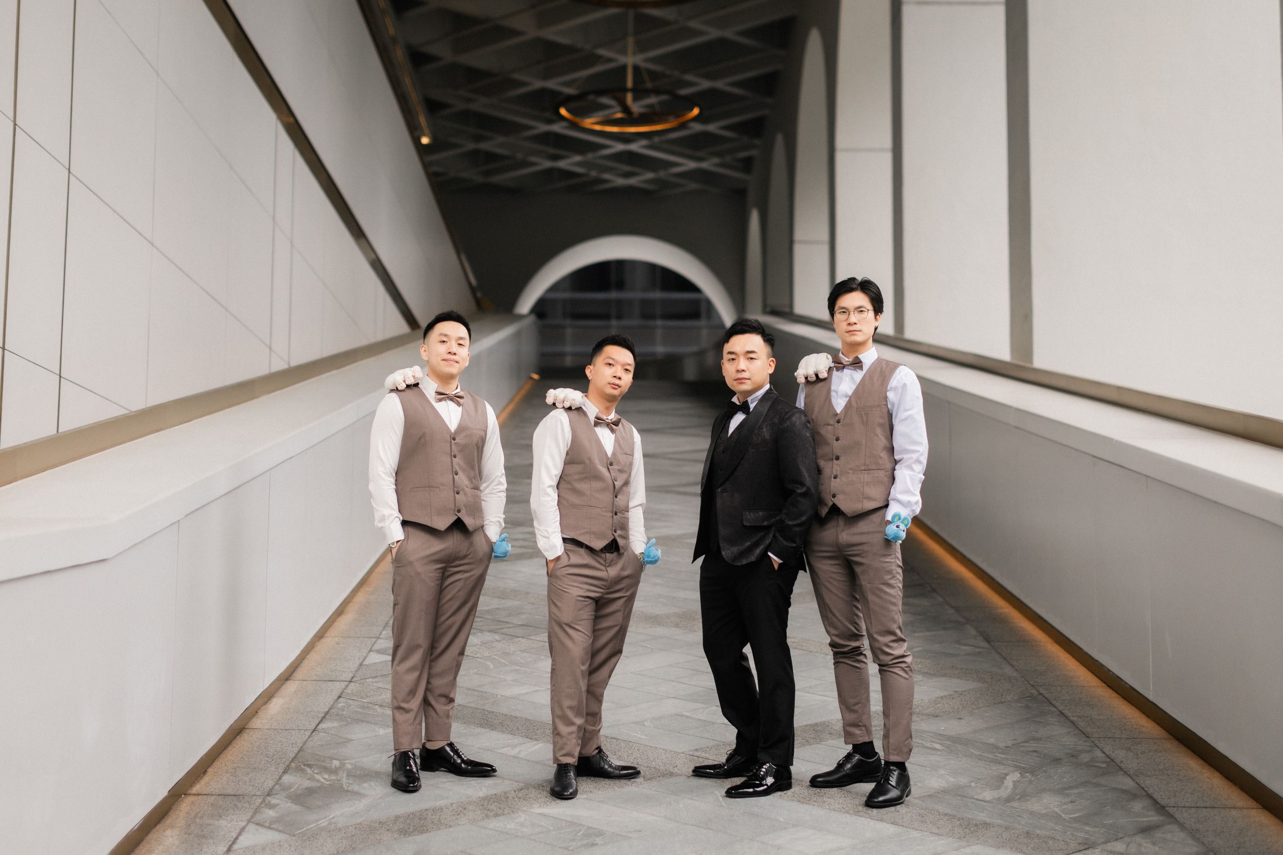 Groom in black suit posing with three groomsmen in tan vests in a modern architectural hallway.