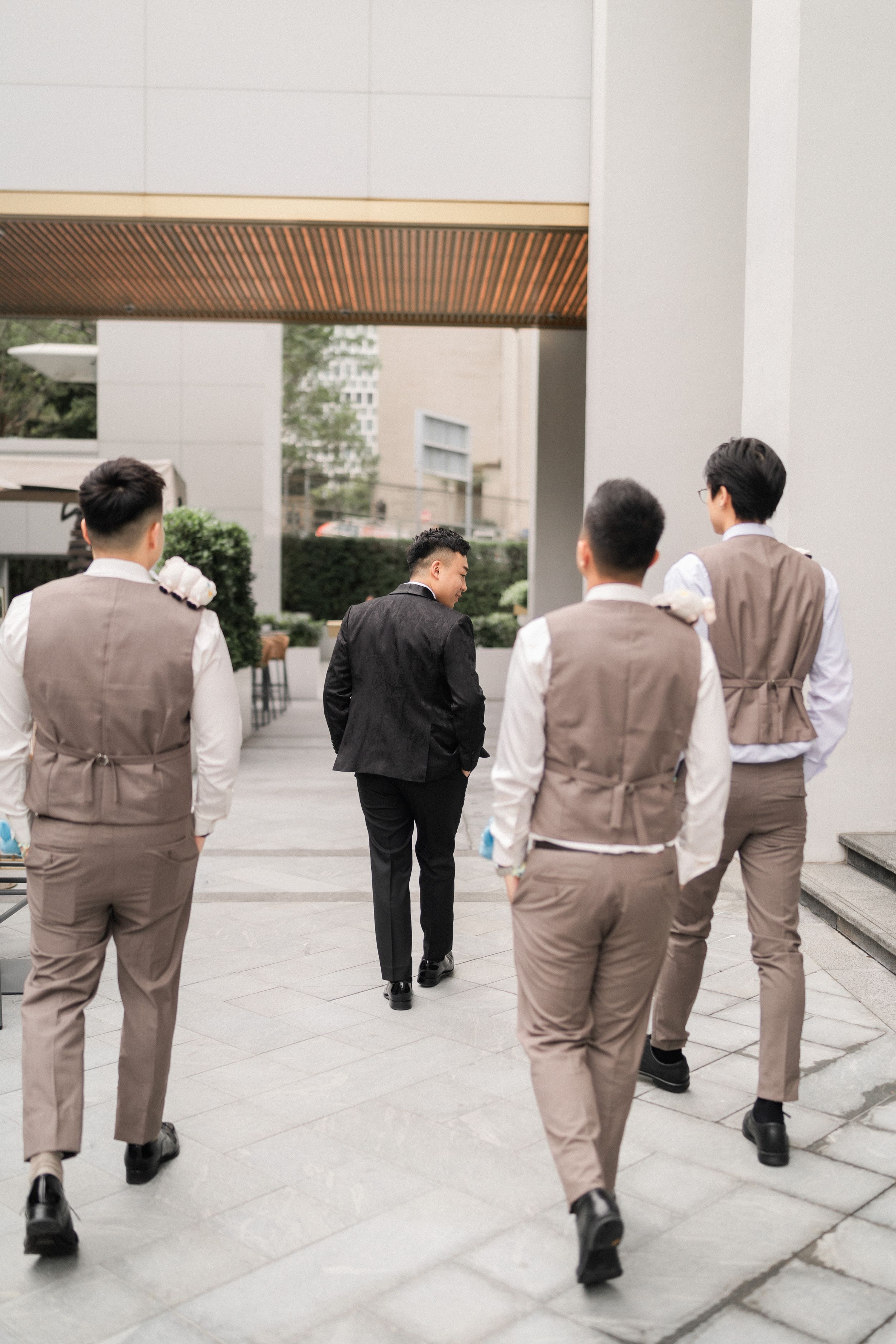 Groomsmen in tan vests and groom in black suit walking through modern outdoor venue.