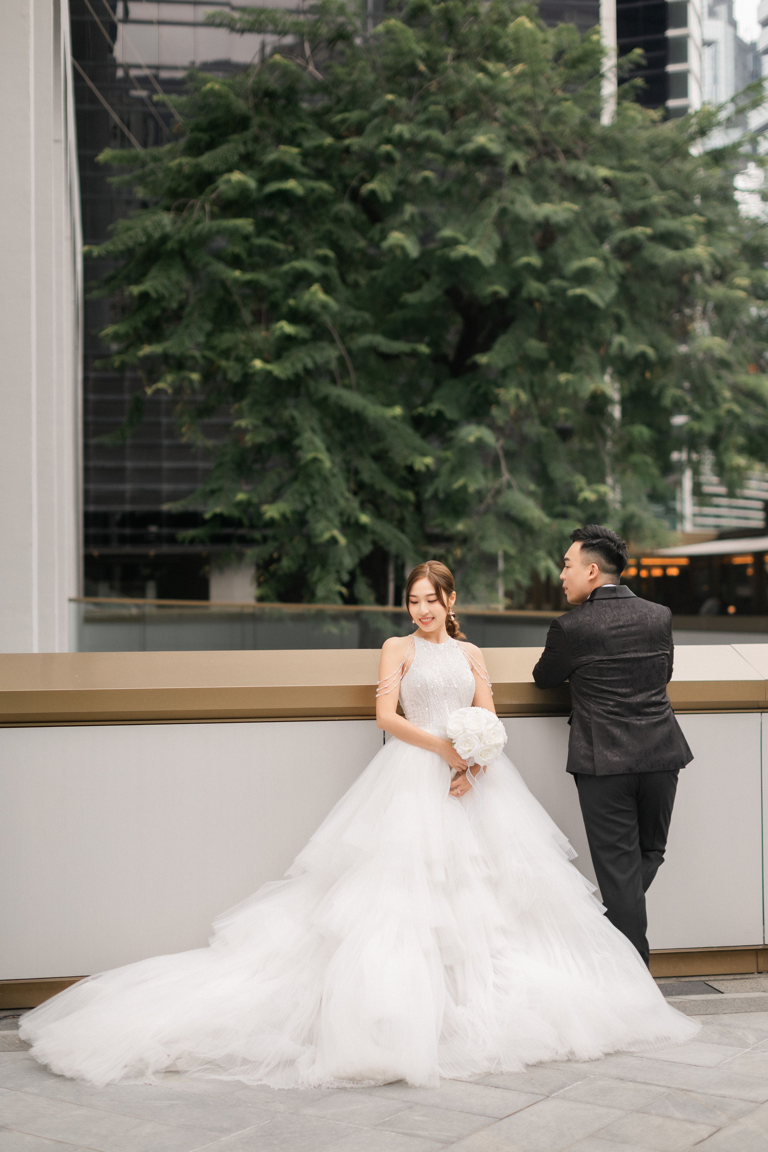 Bride in layered white gown holding bouquet with groom in black suit at outdoor venue.
