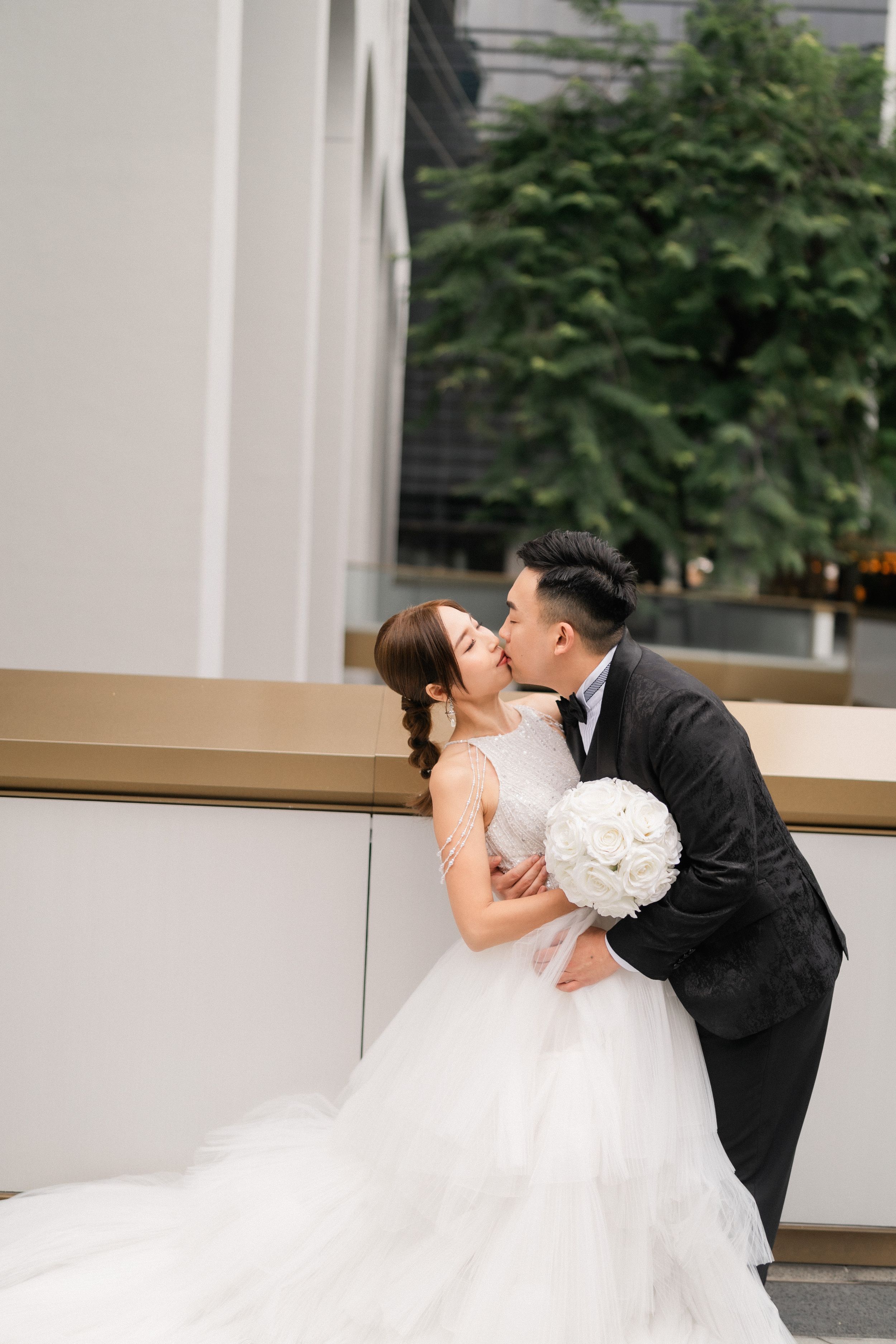 Groom kissing bride in elegant white tiered wedding gown outdoors with lush greenery.