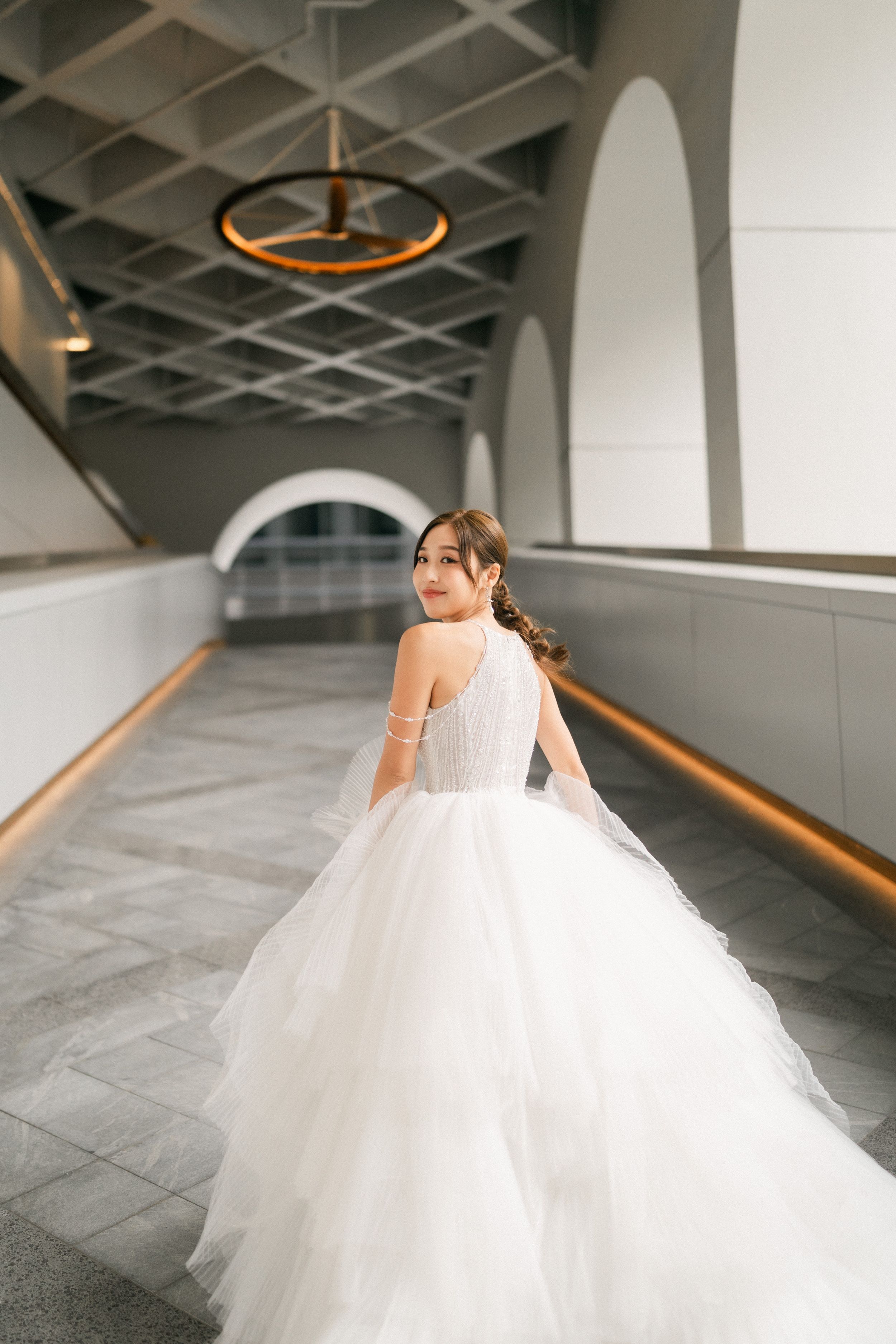 Bride in elegant white tulle ball gown looks over shoulder in modern architectural corridor.