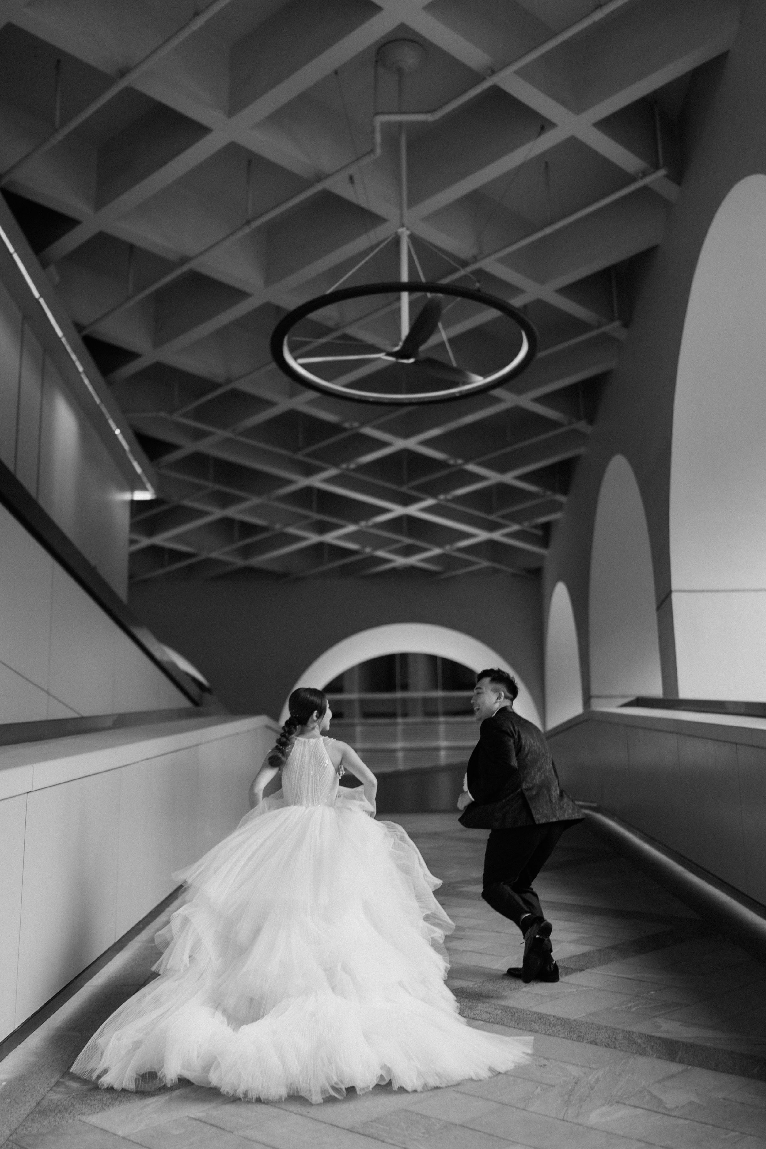 Black and white wedding photo of couple walking in modern hallway with geometric ceiling details.