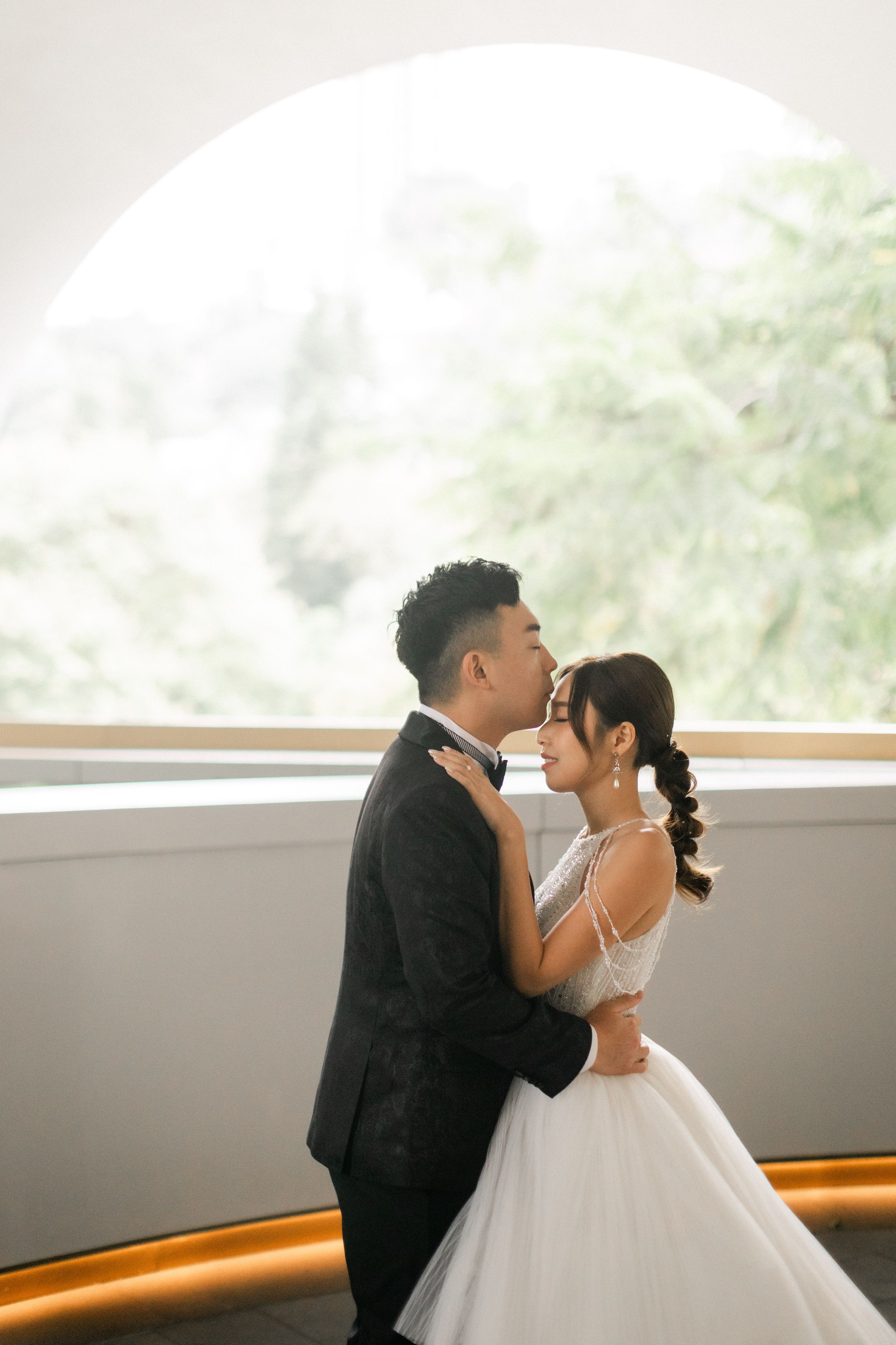 Groom in black suit kisses bride in white gown near large arched window with greenery backdrop.
