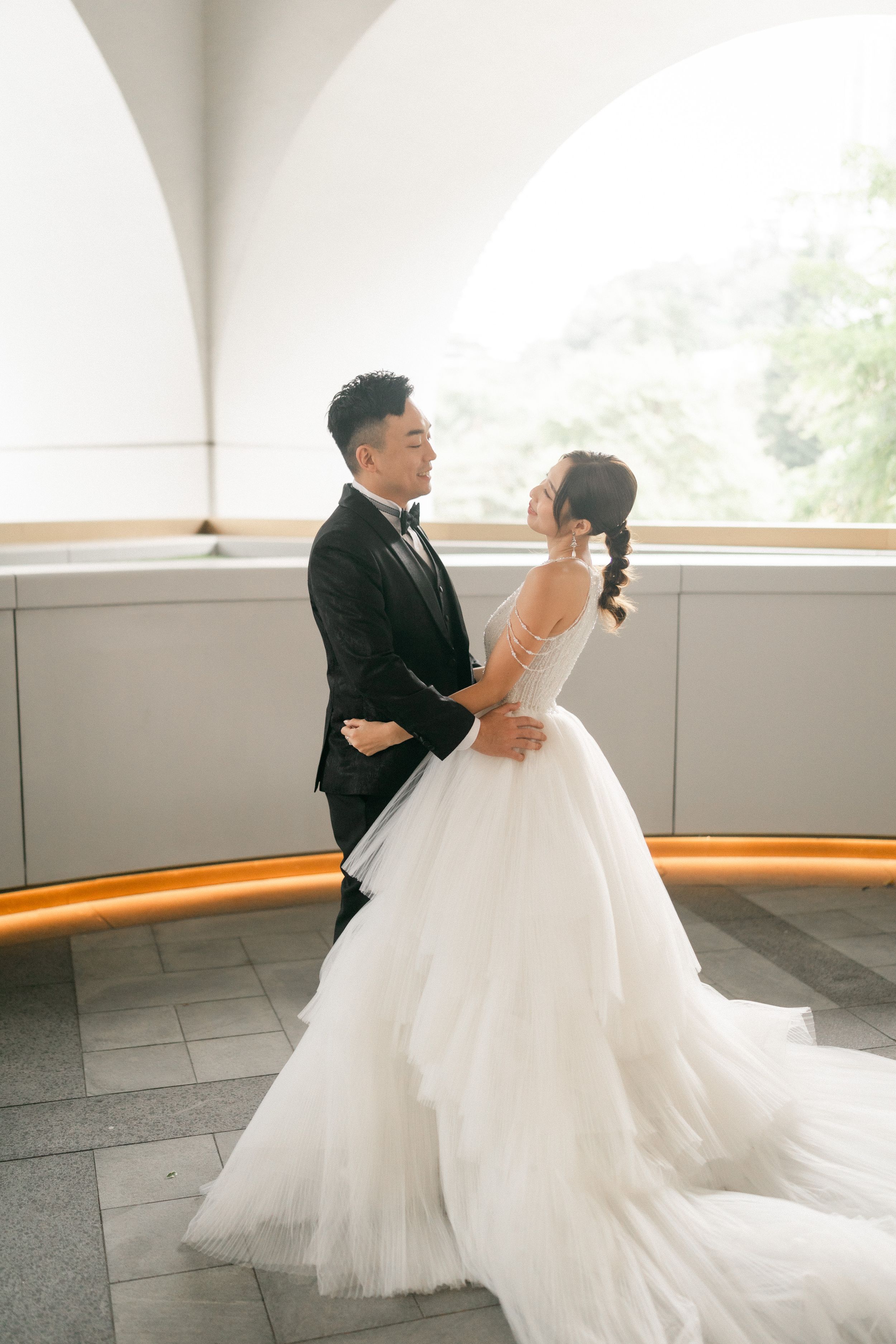 Bride and groom embrace tenderly in bright corridor with arched windows and white tulle gown.
