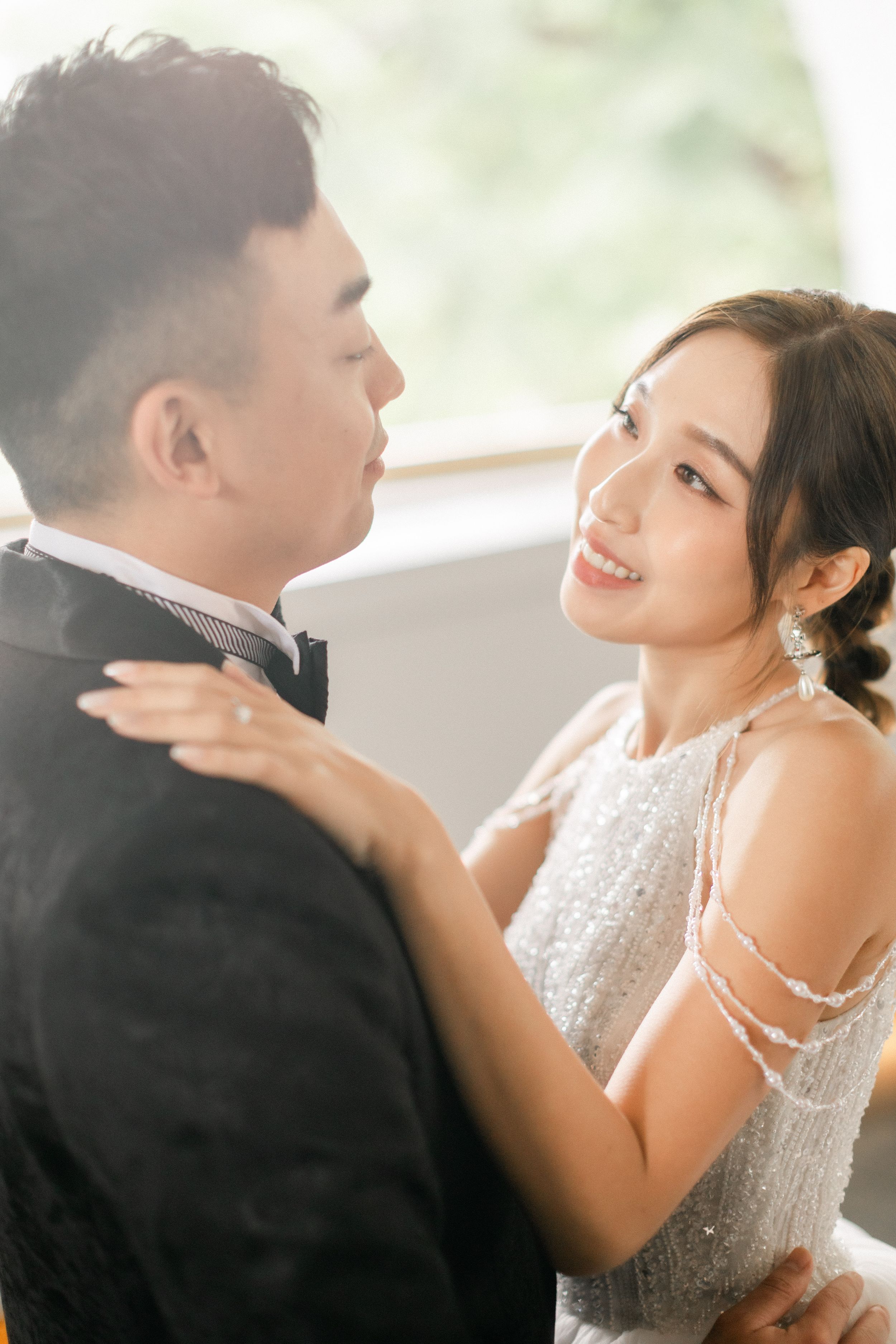 Smiling bride in silver beaded gown gazes at groom in black tuxedo in soft natural light.