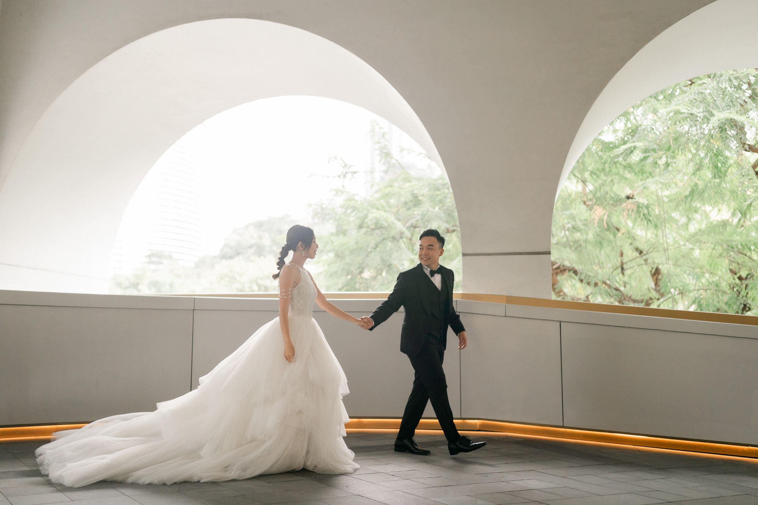 Bride and groom hold hands walking through elegant white arched corridor with lush green trees visible through windows.