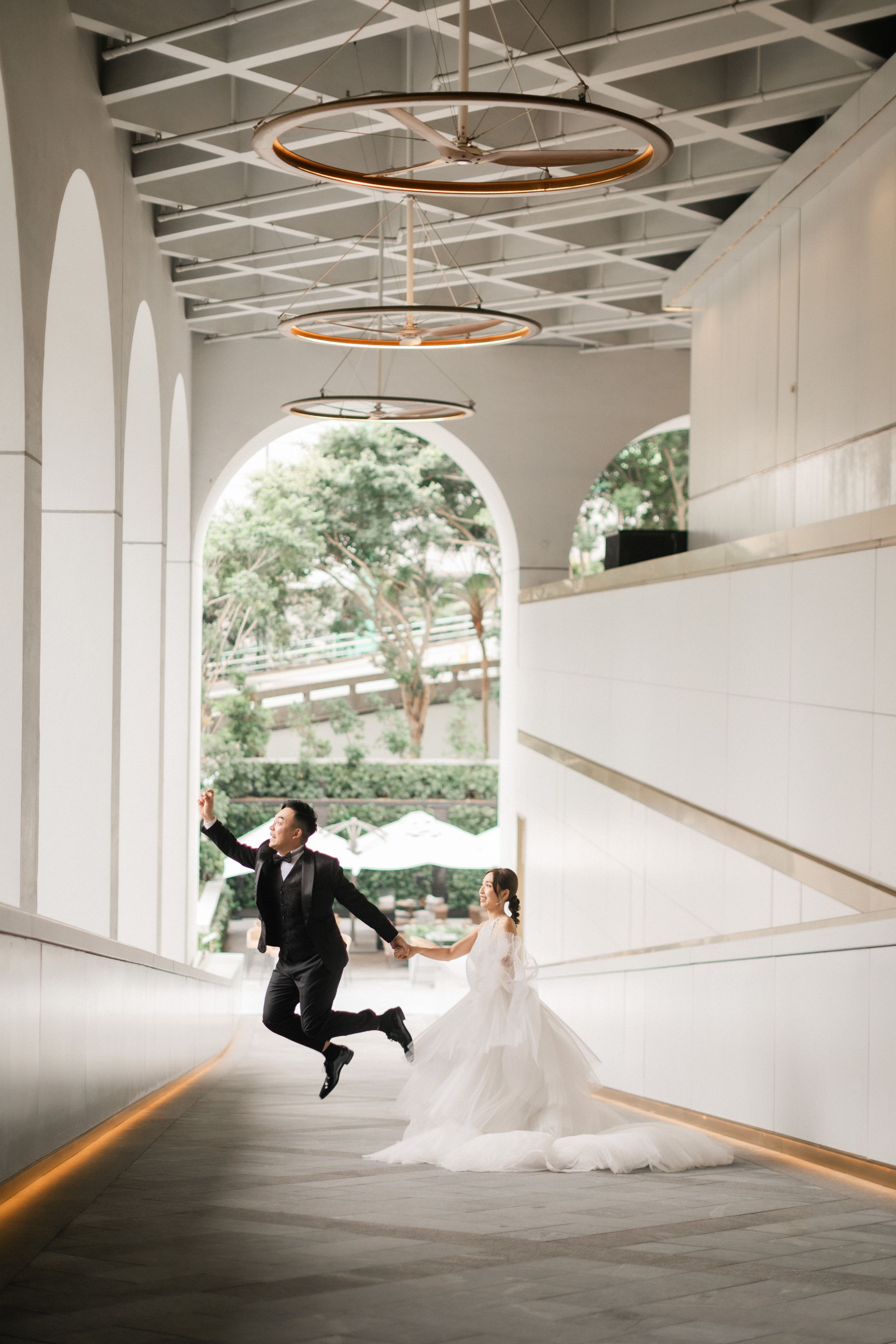 Joyful groom jumps mid-air holding brides hand under circular chandelier in modern white arched wedding venue.