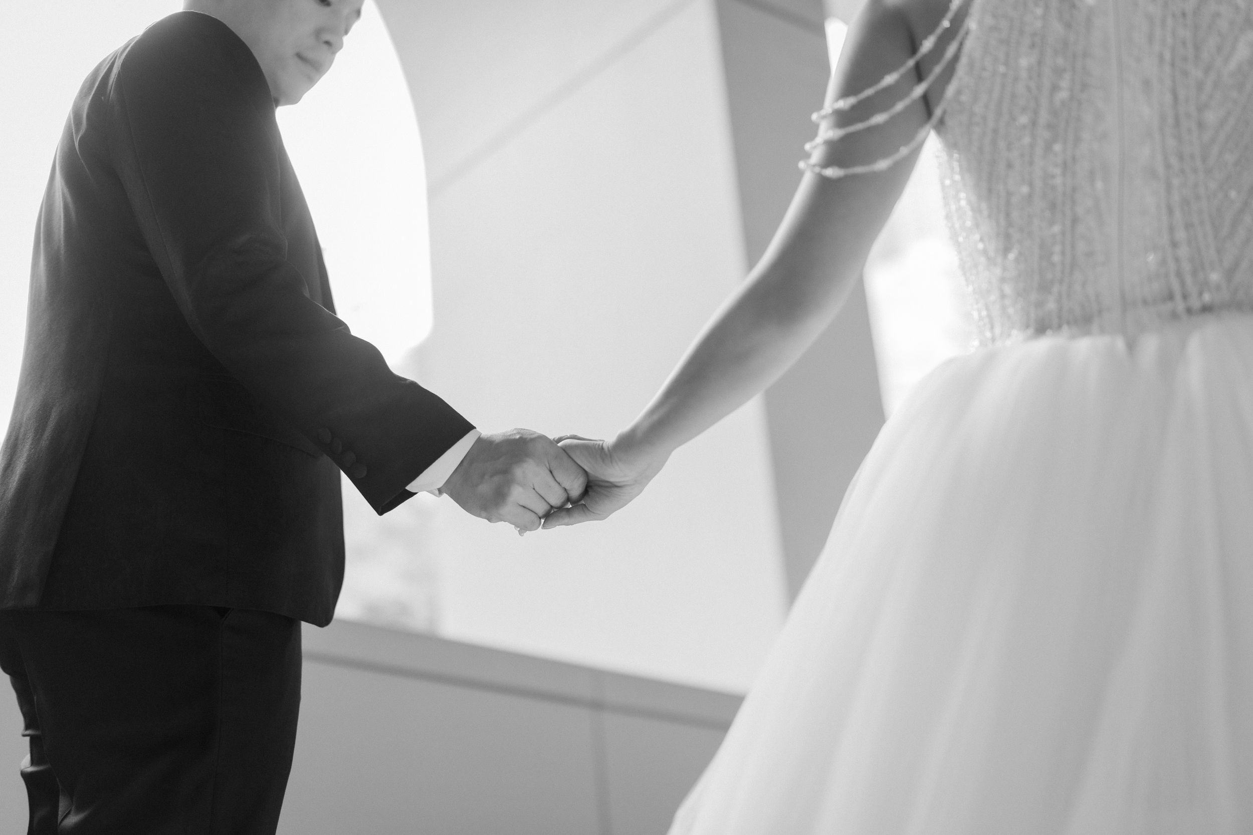 Black and white close-up of bride and groom holding hands tenderly in soft natural light.