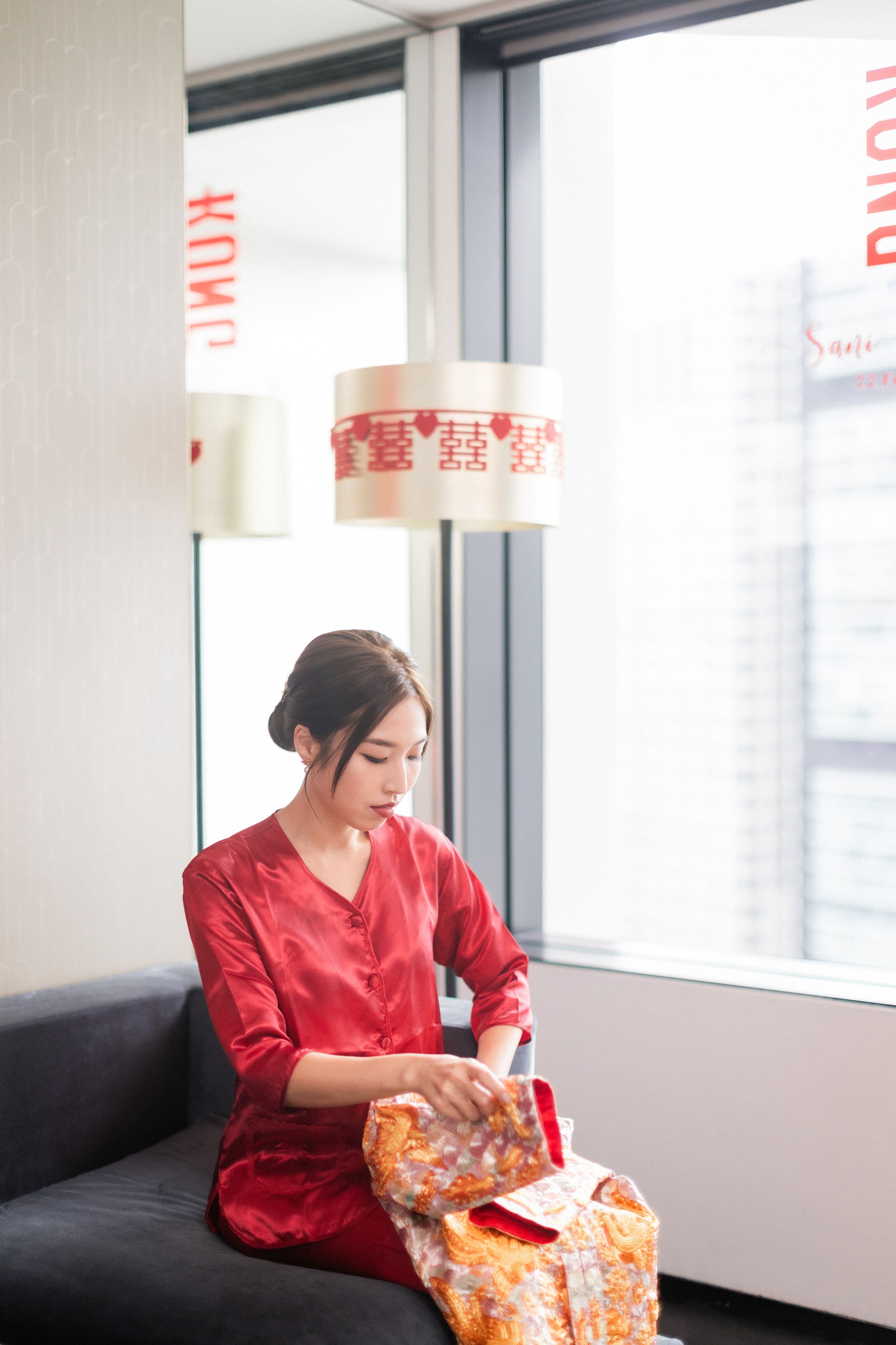 Bride in red silk robe preparing for wedding ceremony seated by window with double happiness decor.