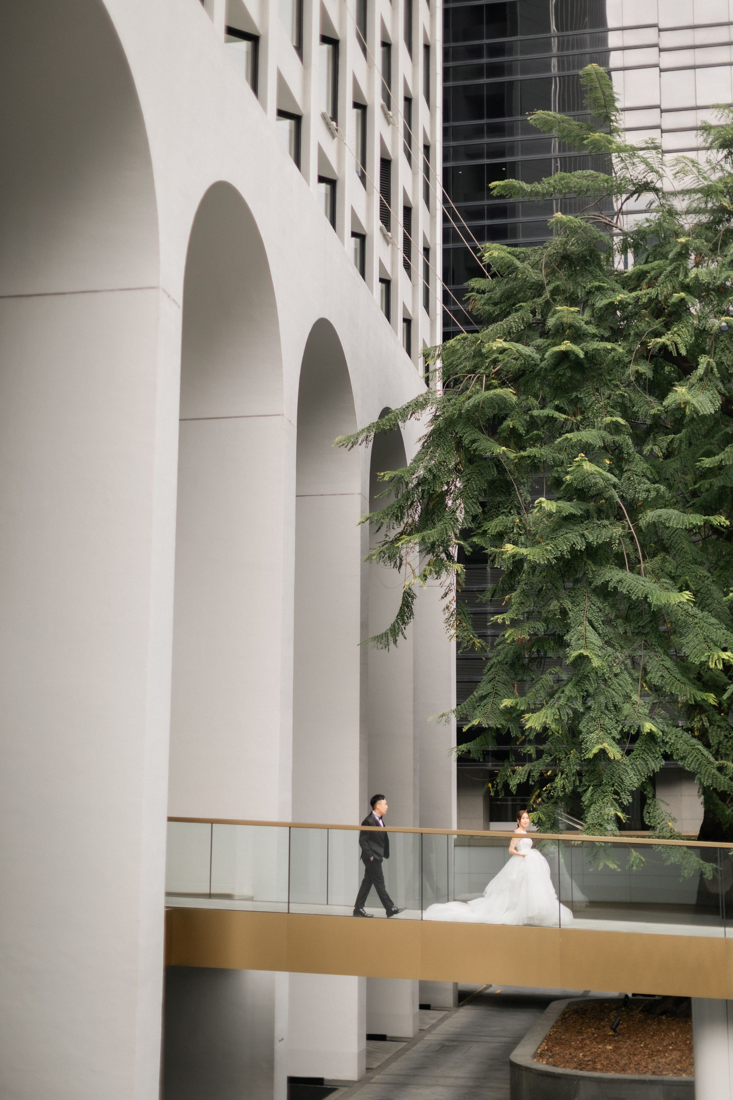 Bride in flowing white gown and groom walk along modern architectural corridor with tall arches and green trees.