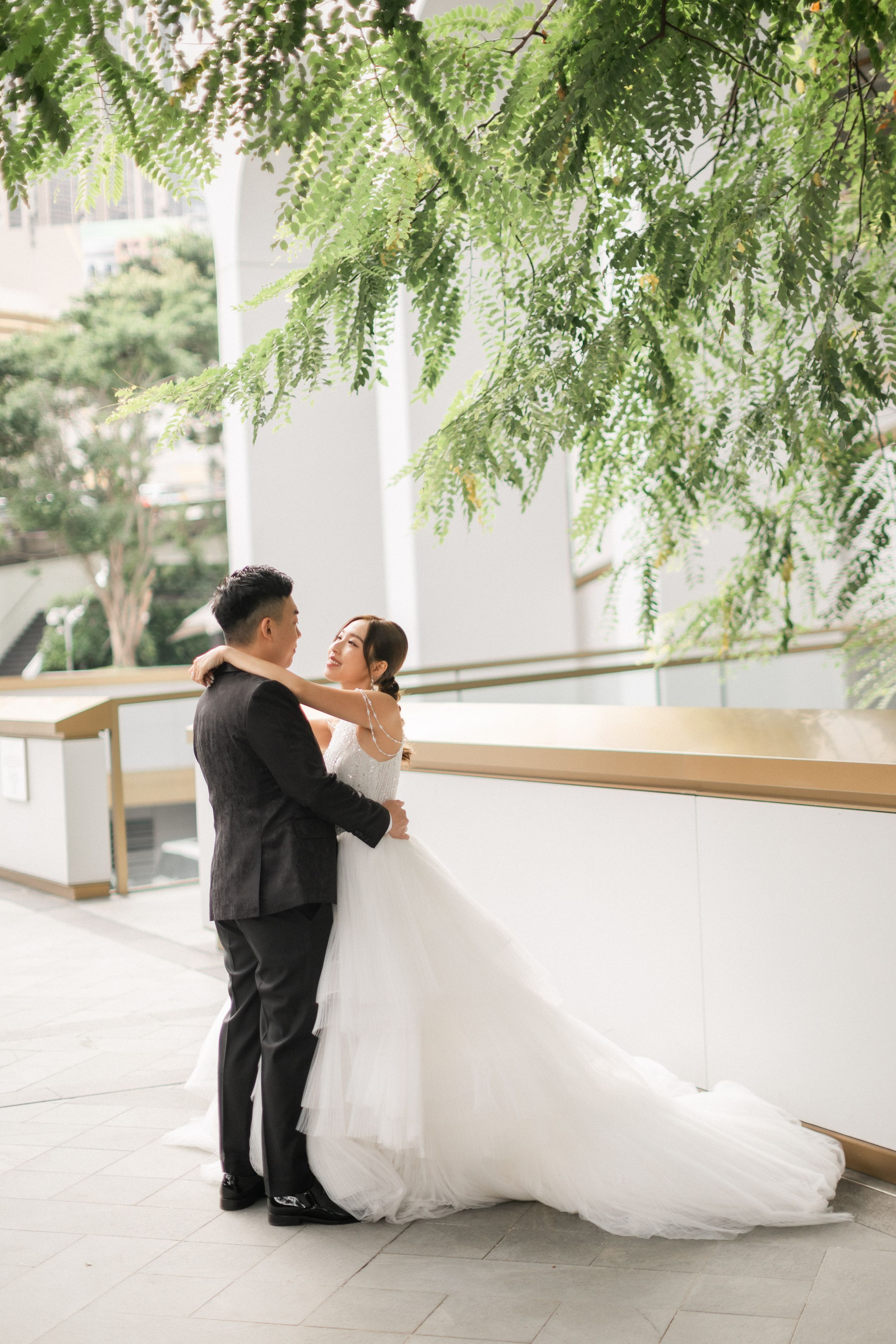 Bride and groom embrace romantically under hanging green ferns in elegant outdoor wedding venue setting.