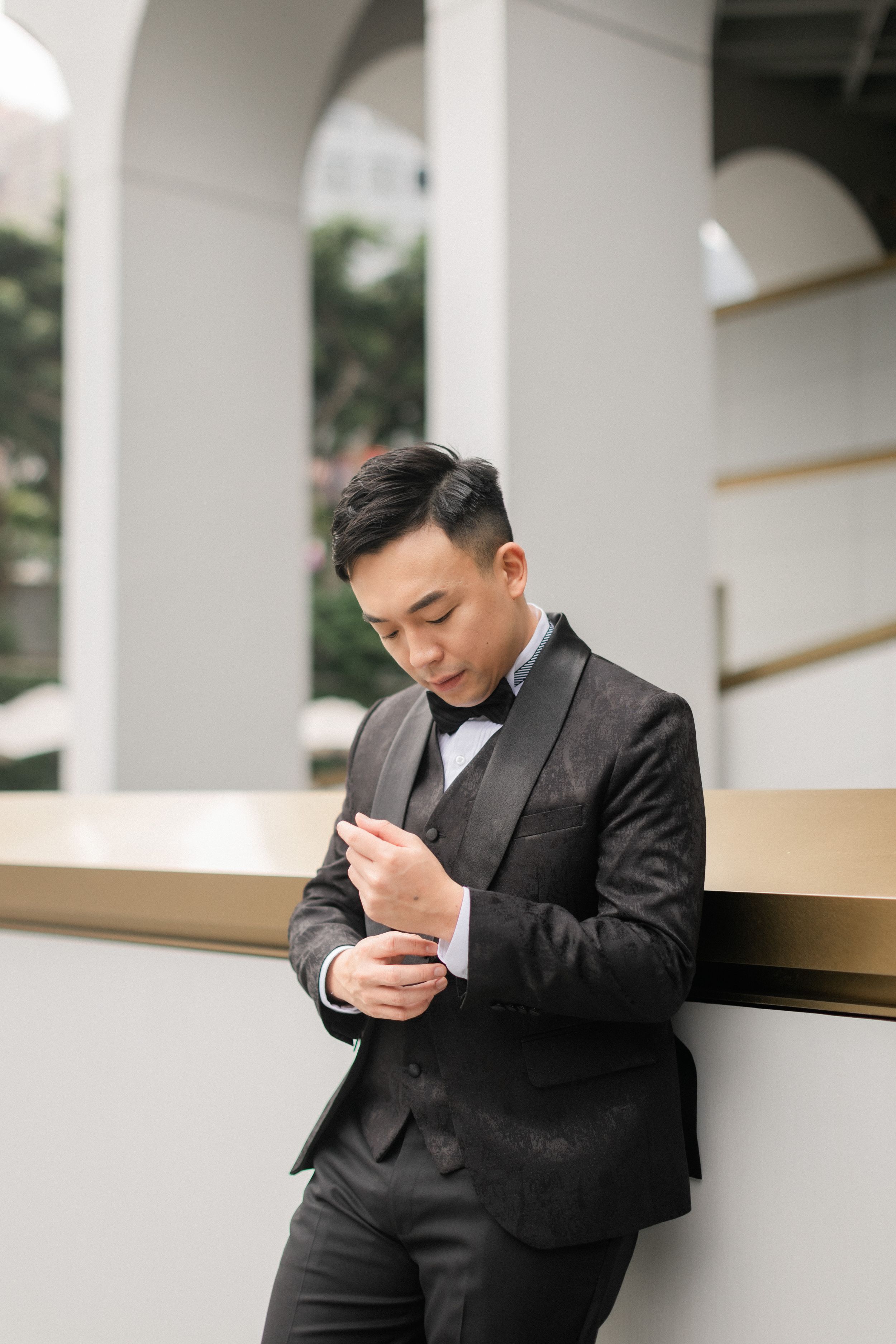 Dapper groom in black tuxedo adjusts cufflinks while leaning against gold railing in white arched venue.