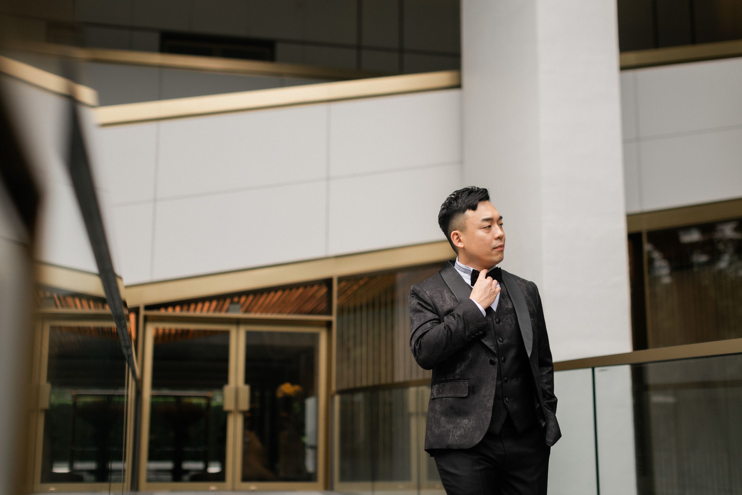 Stylish groom in dark suit adjusts bow tie outdoors near modern glass building entrance.