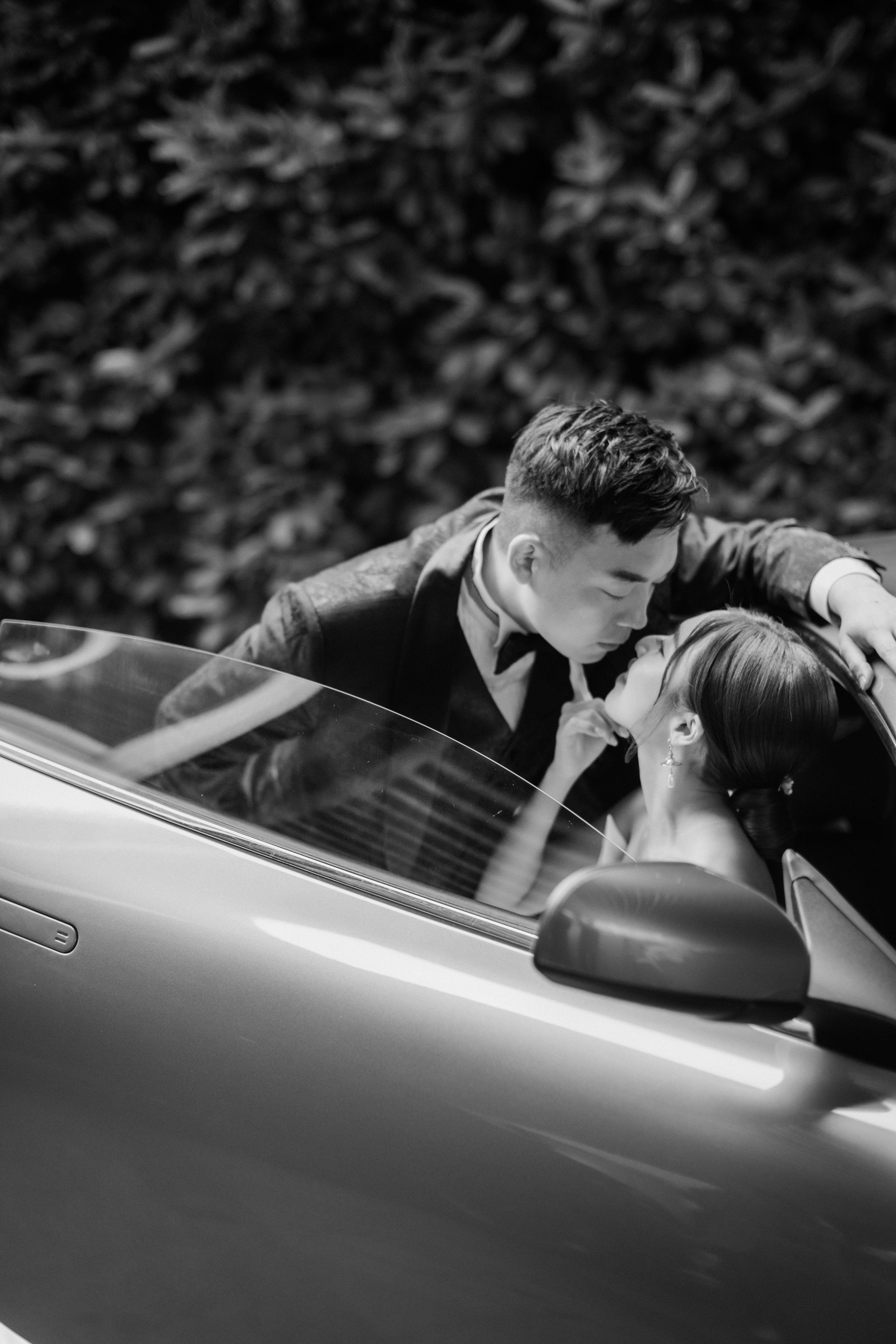 Black and white romantic photo of bride and groom sharing a kiss leaning over a luxury convertible car.