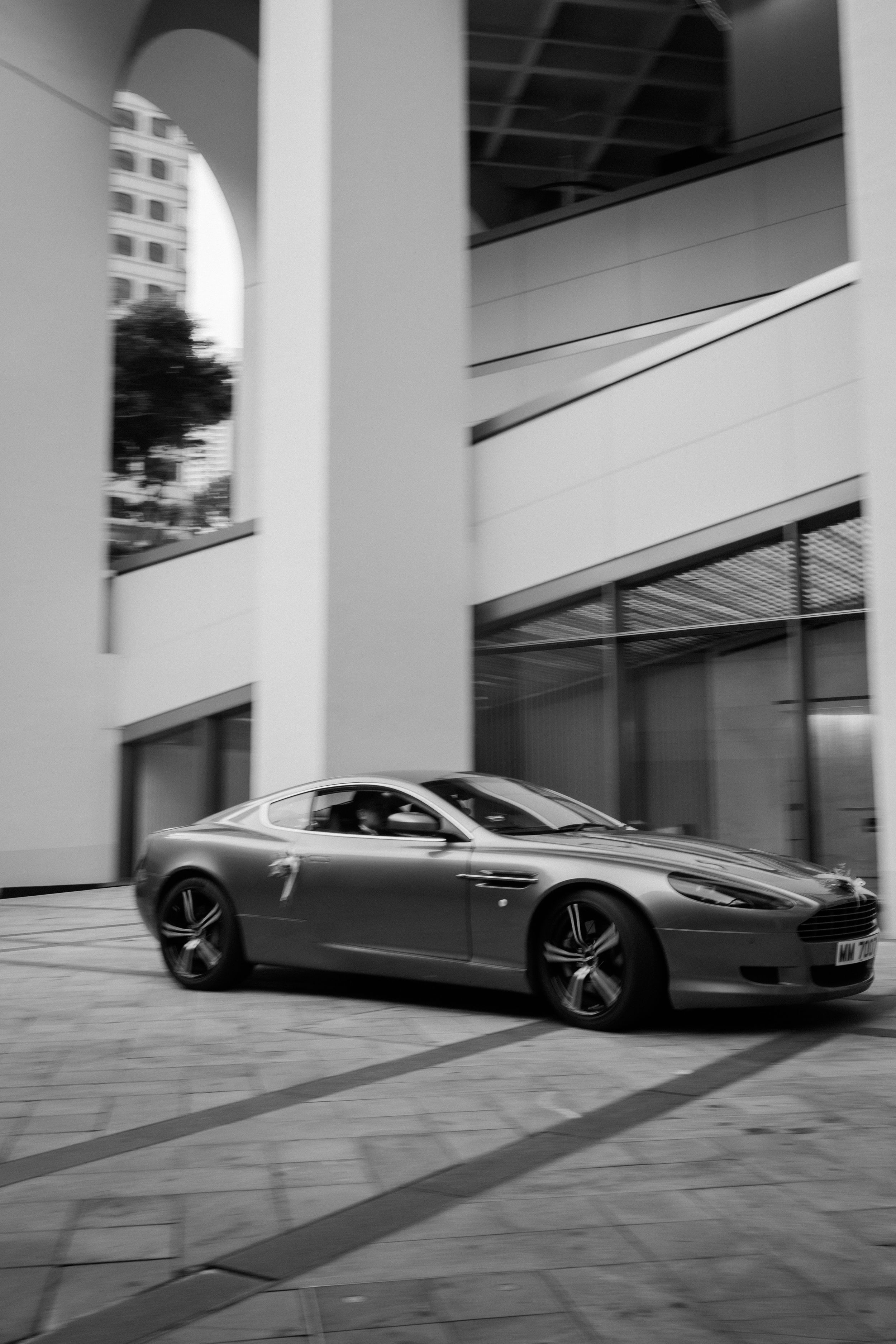 Black and white photo of an Aston Martin DB9 parked beside a modern architectural building.