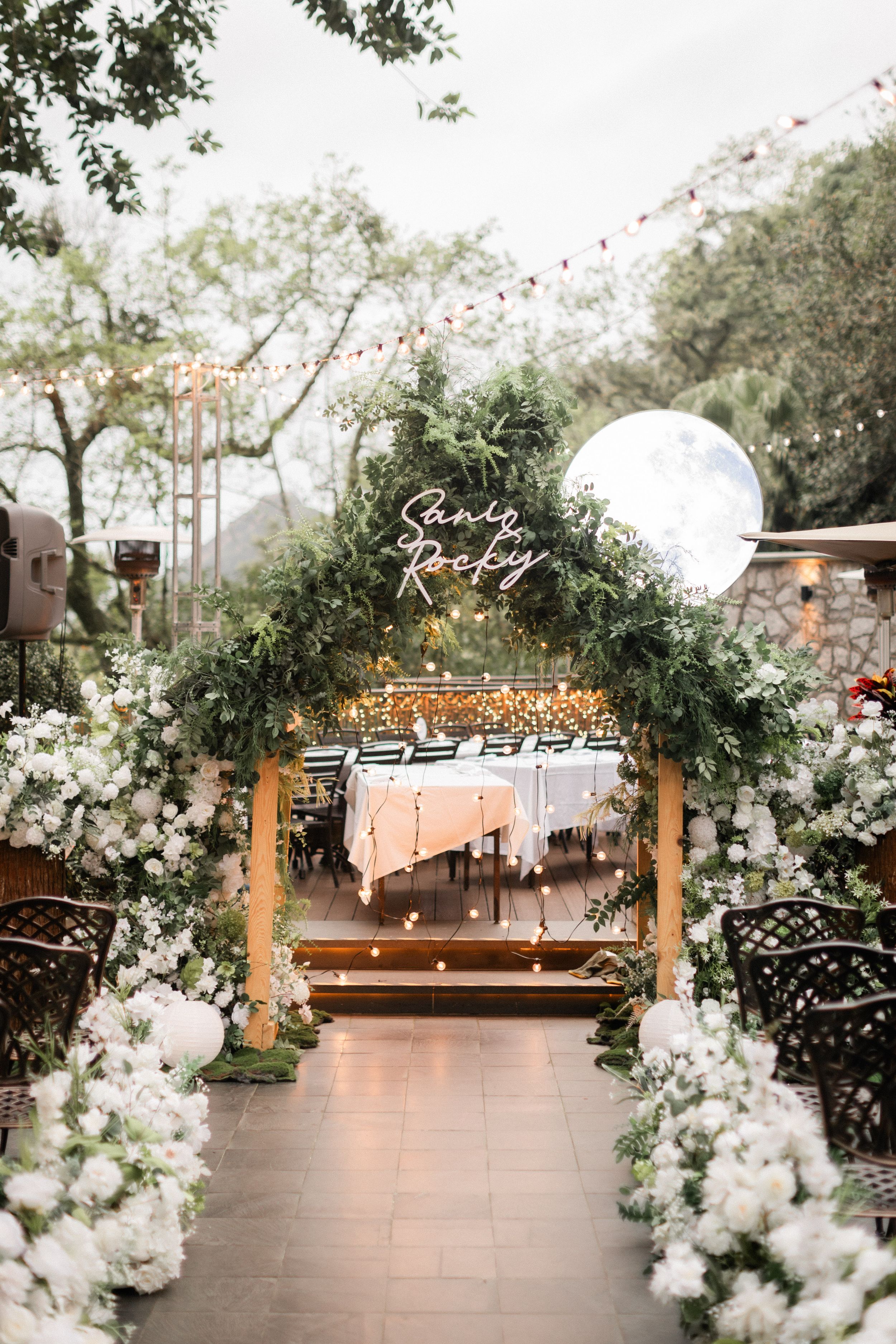 Elegant outdoor wedding arch adorned with lush greenery and white flowers with neon sign reading Sandy Rocky.