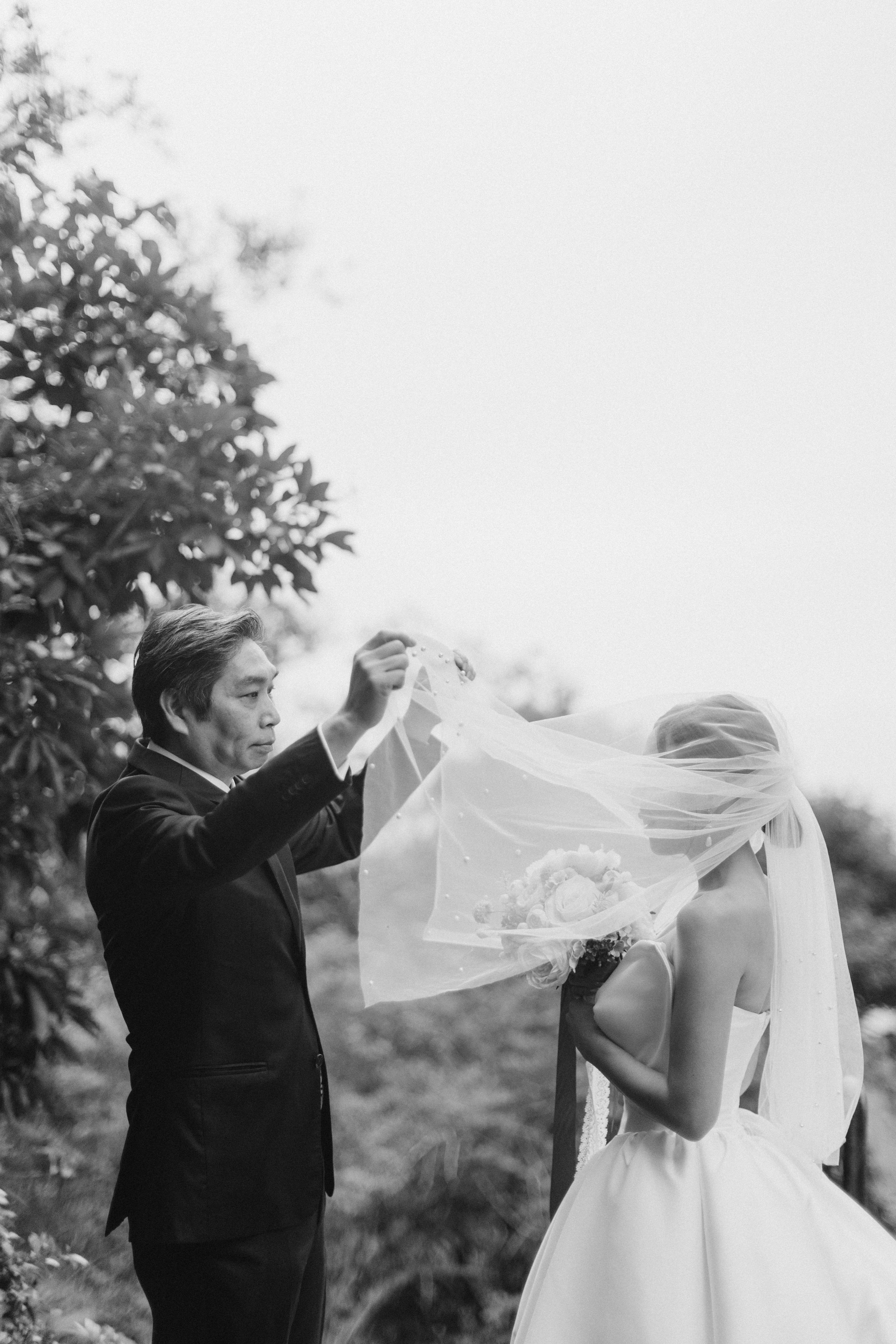 Black and white photo of groom lifting bride's veil outdoors surrounded by lush trees.