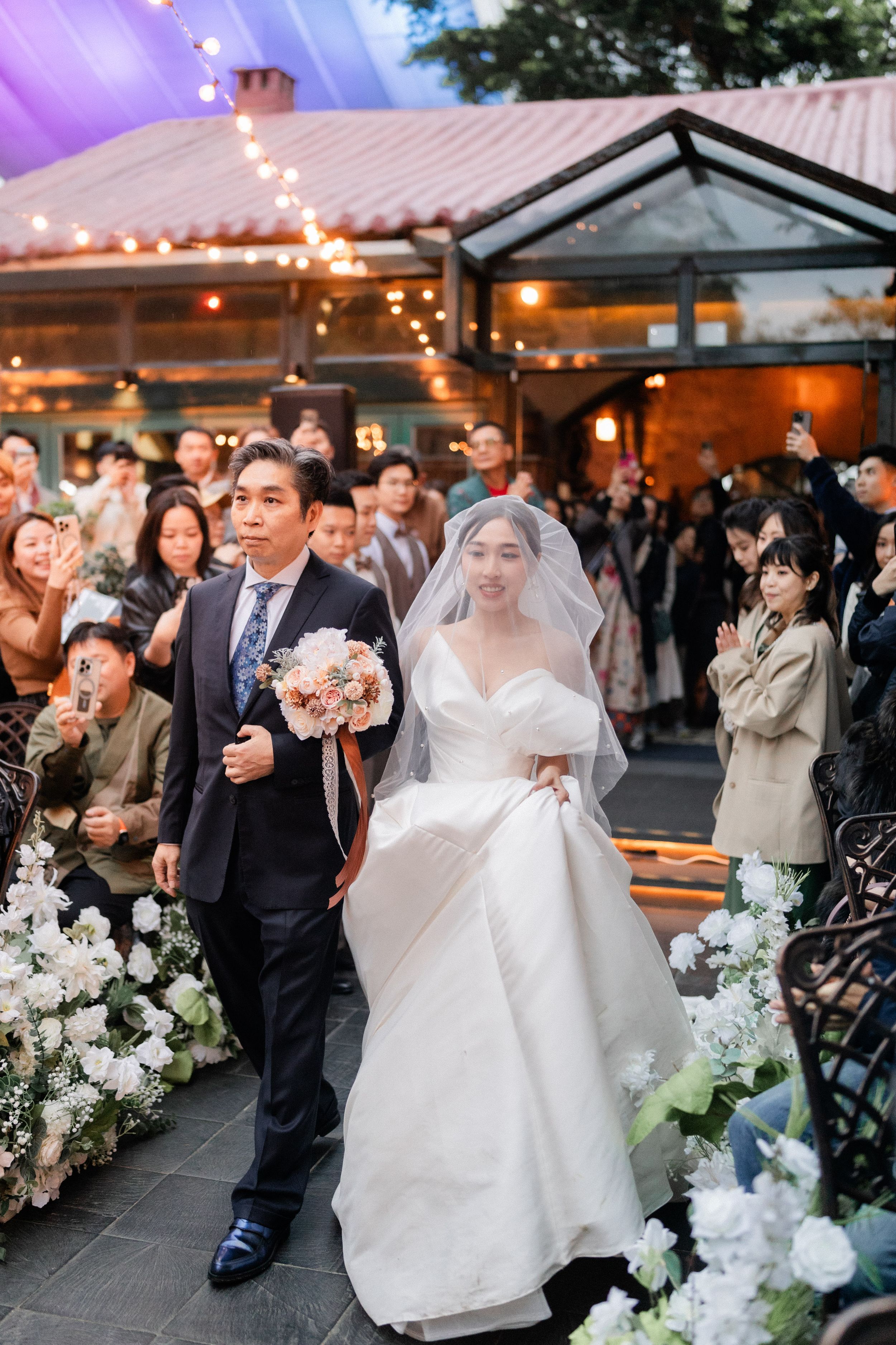 Bride in a white gown carrying a bouquet walks down the aisle with her escort at an outdoor wedding.