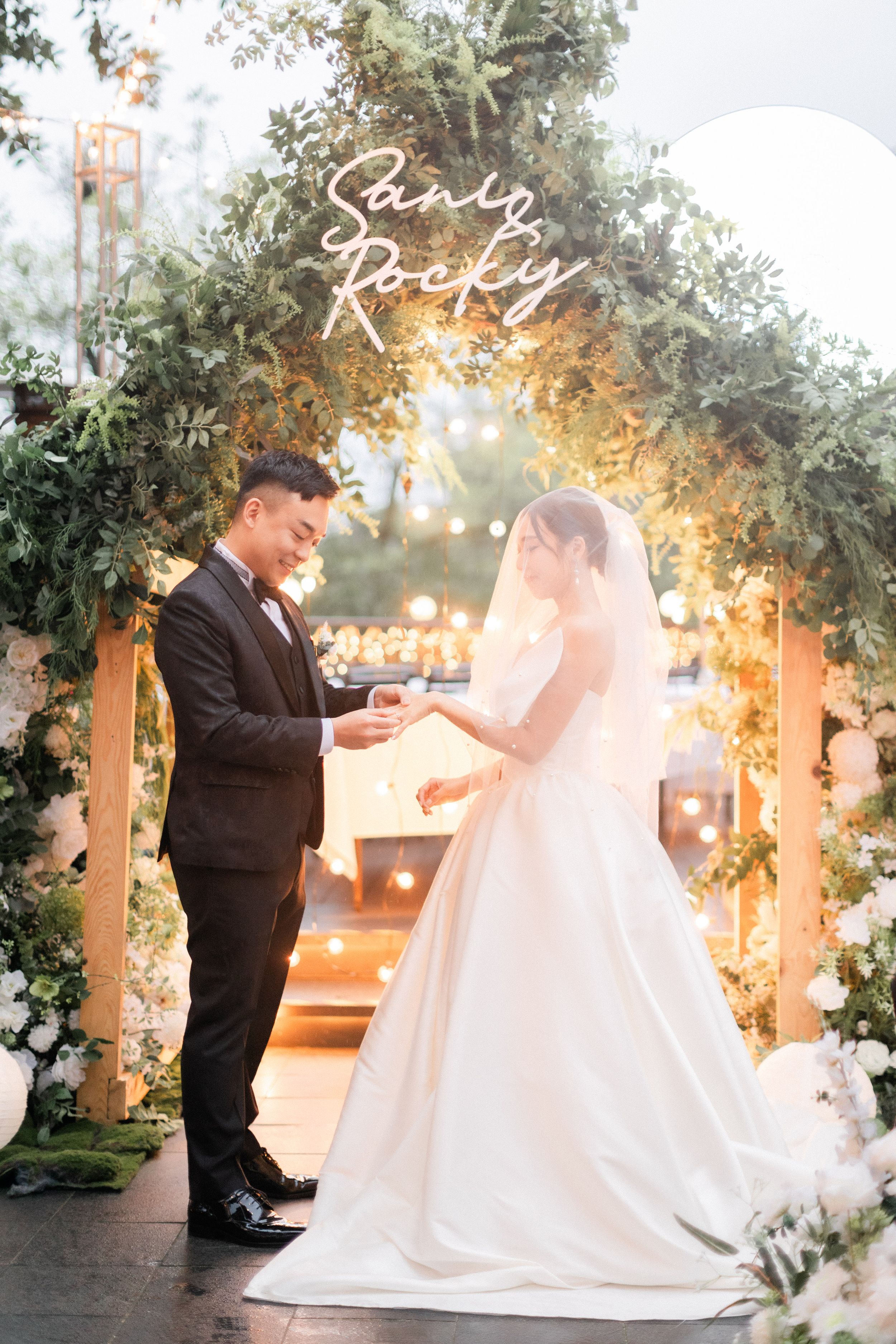Bride and groom exchanging rings under a floral arch with a neon sign at their wedding.