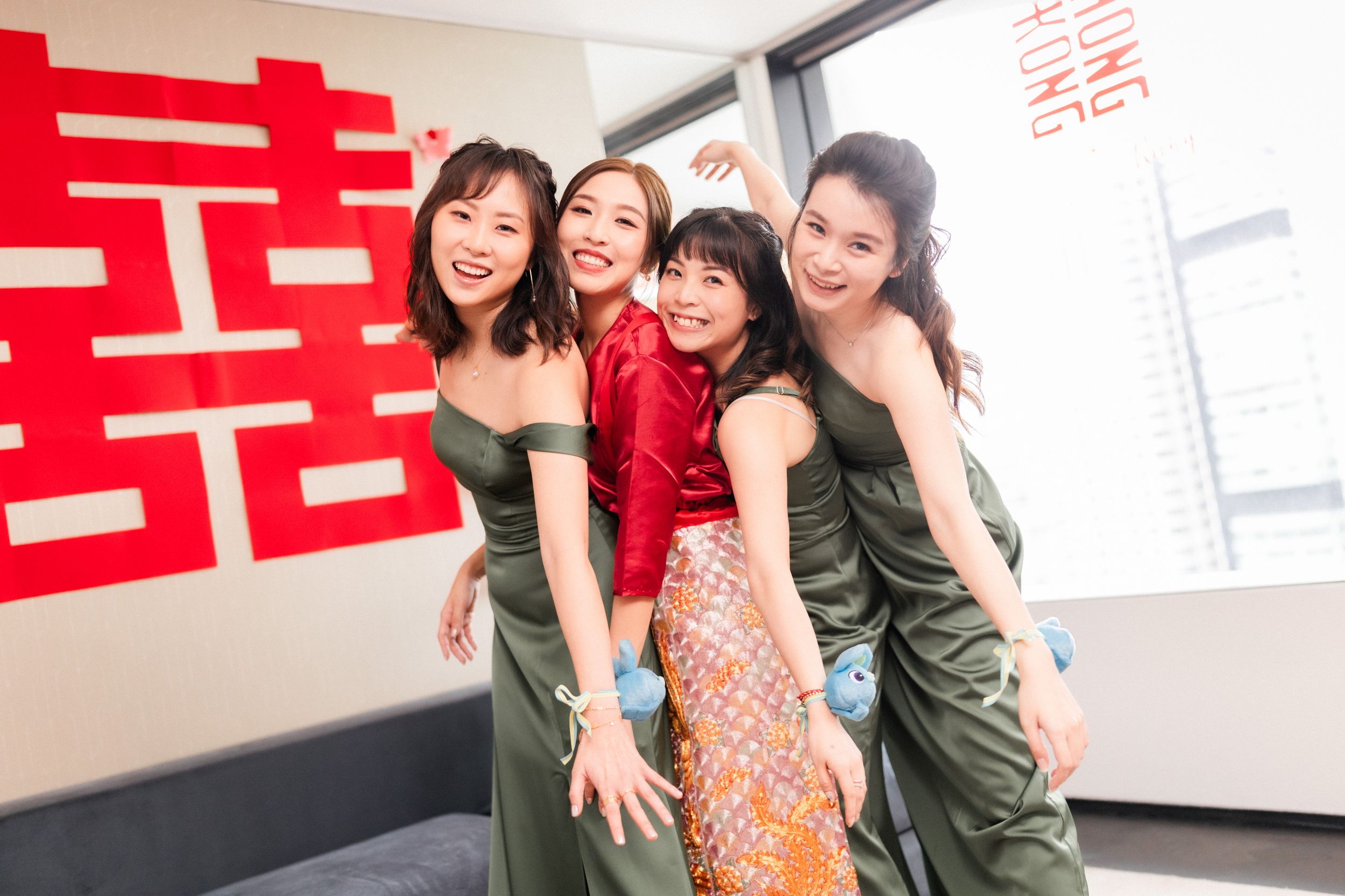 Four women in Chinese wedding attire smile and pose together in front of a red double happiness symbol.