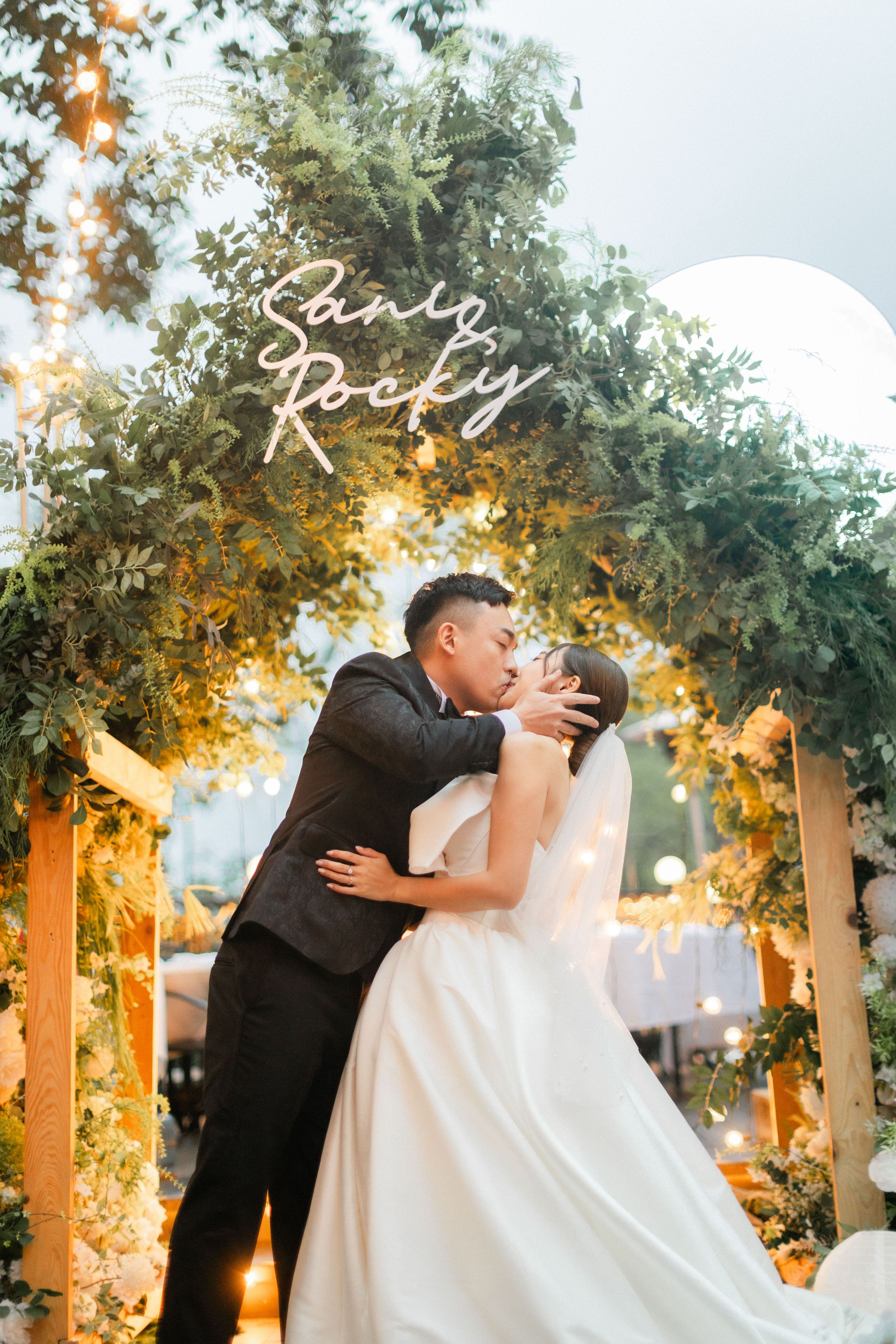 Bride and groom share a romantic kiss under a floral arch with a glowing neon sign at their wedding.