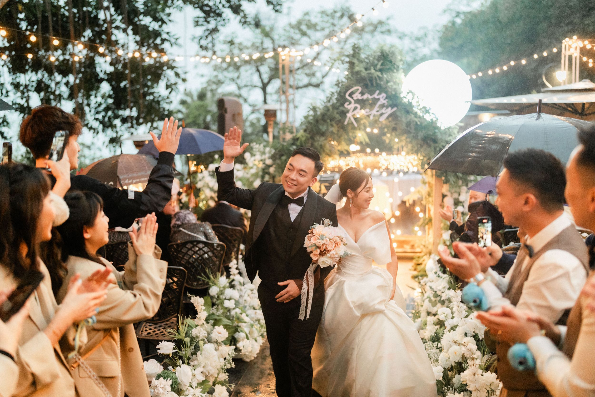 Happy bride and groom walk down the aisle as guests celebrate at an outdoor evening wedding ceremony.