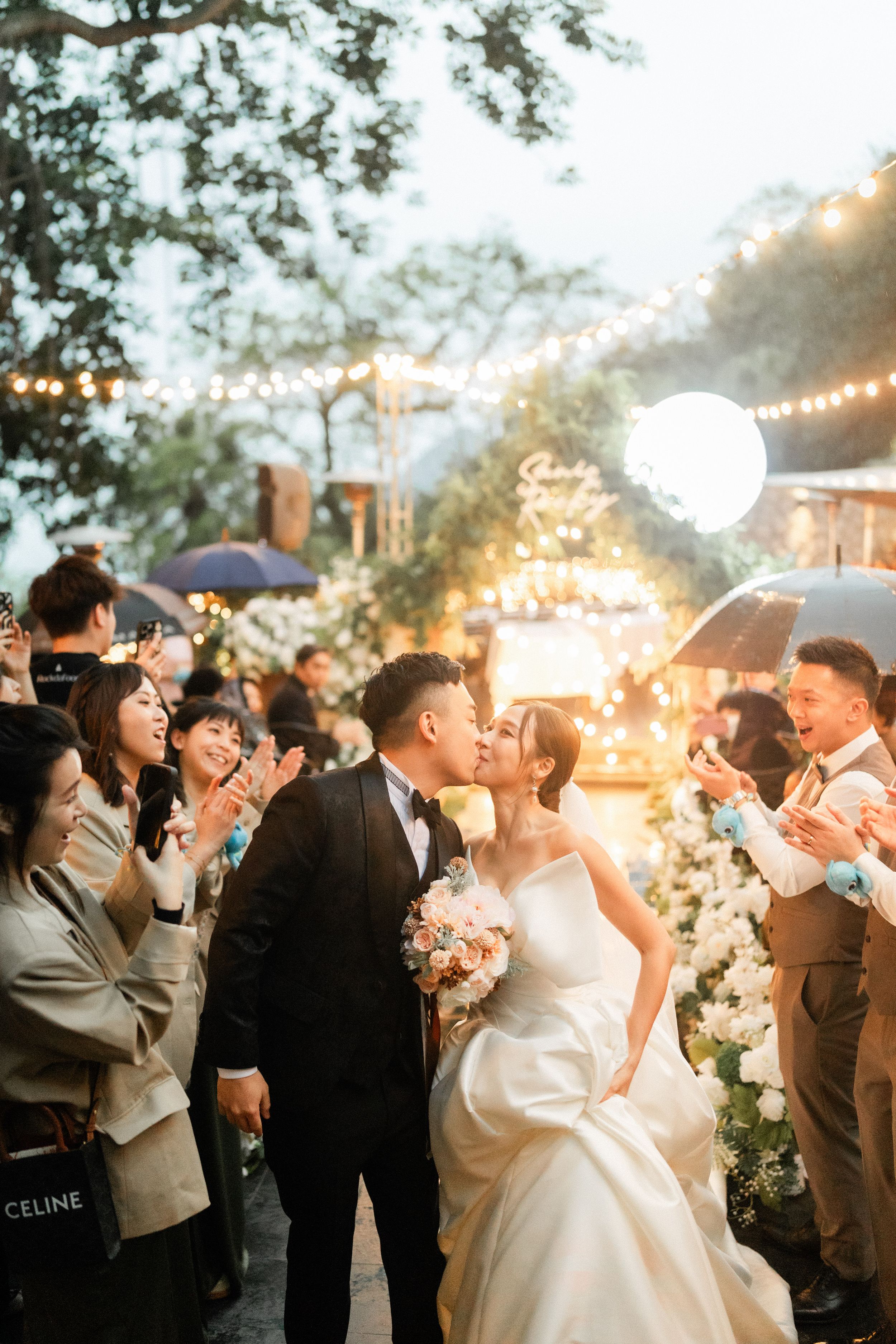 Bride and groom share a kiss while guests cheer and applaud at a beautifully lit outdoor wedding.