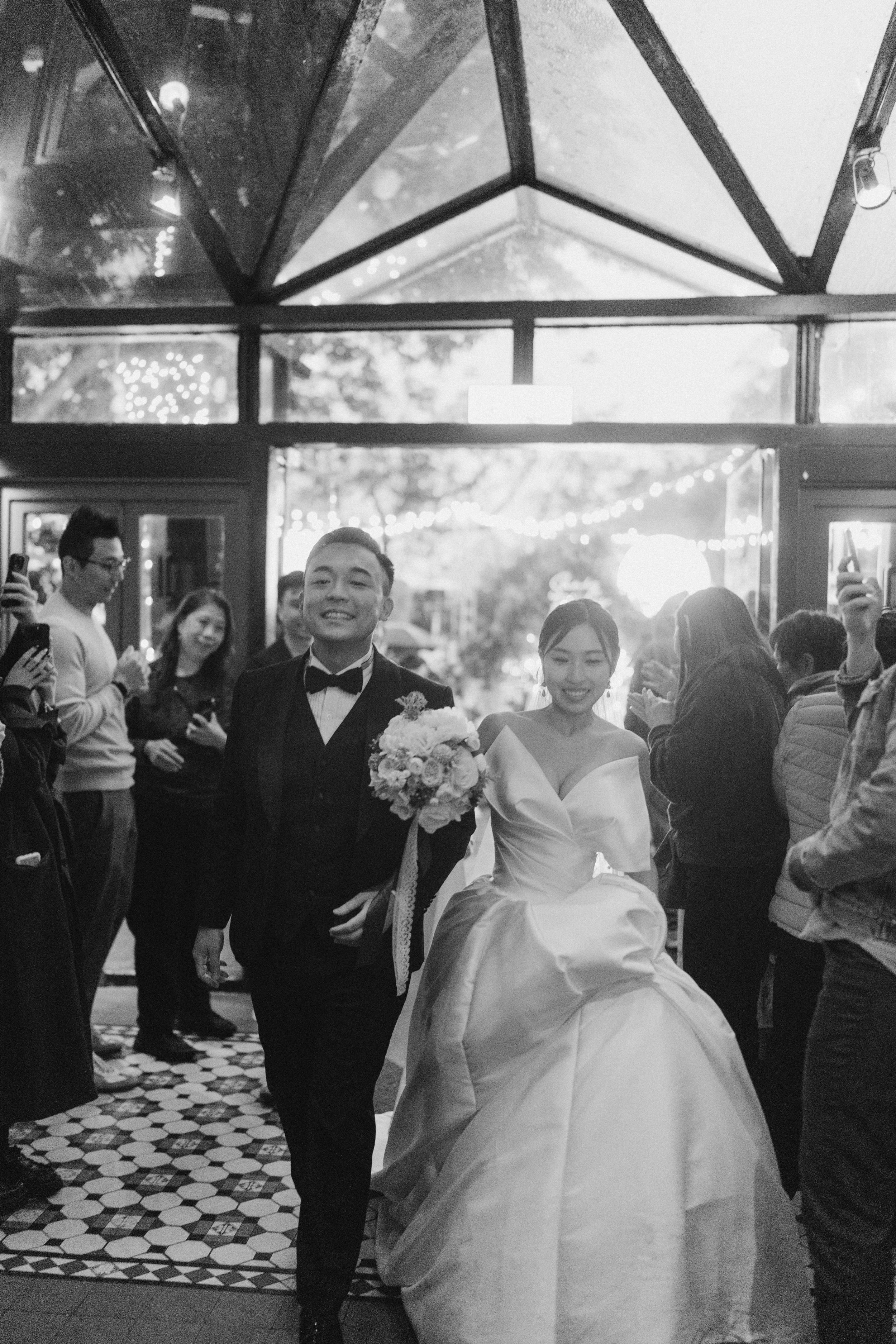 Bride and groom smile as they walk through a doorway surrounded by guests in a black and white photo.