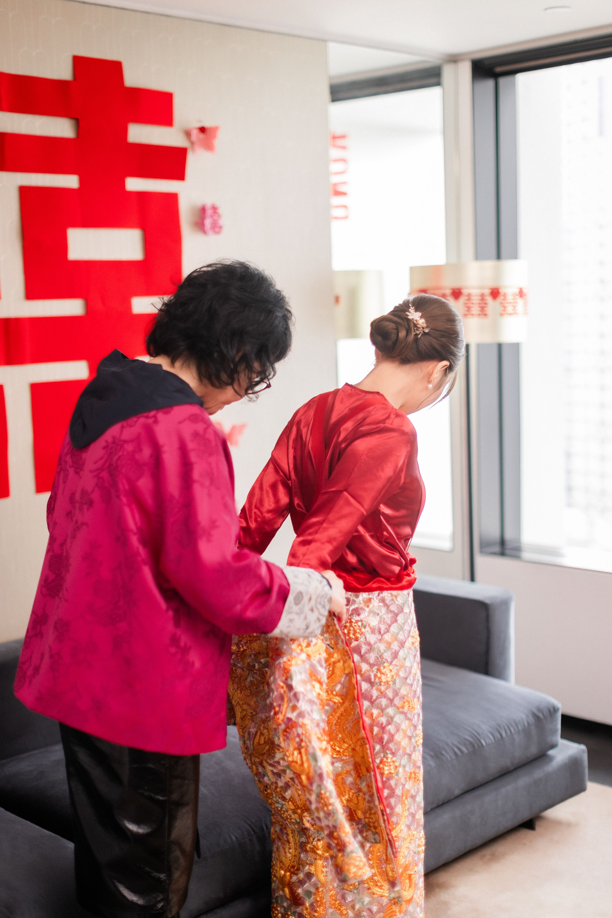 An older woman in pink helps a bride in red qipao and floral skirt prepare for a traditional Chinese wedding.