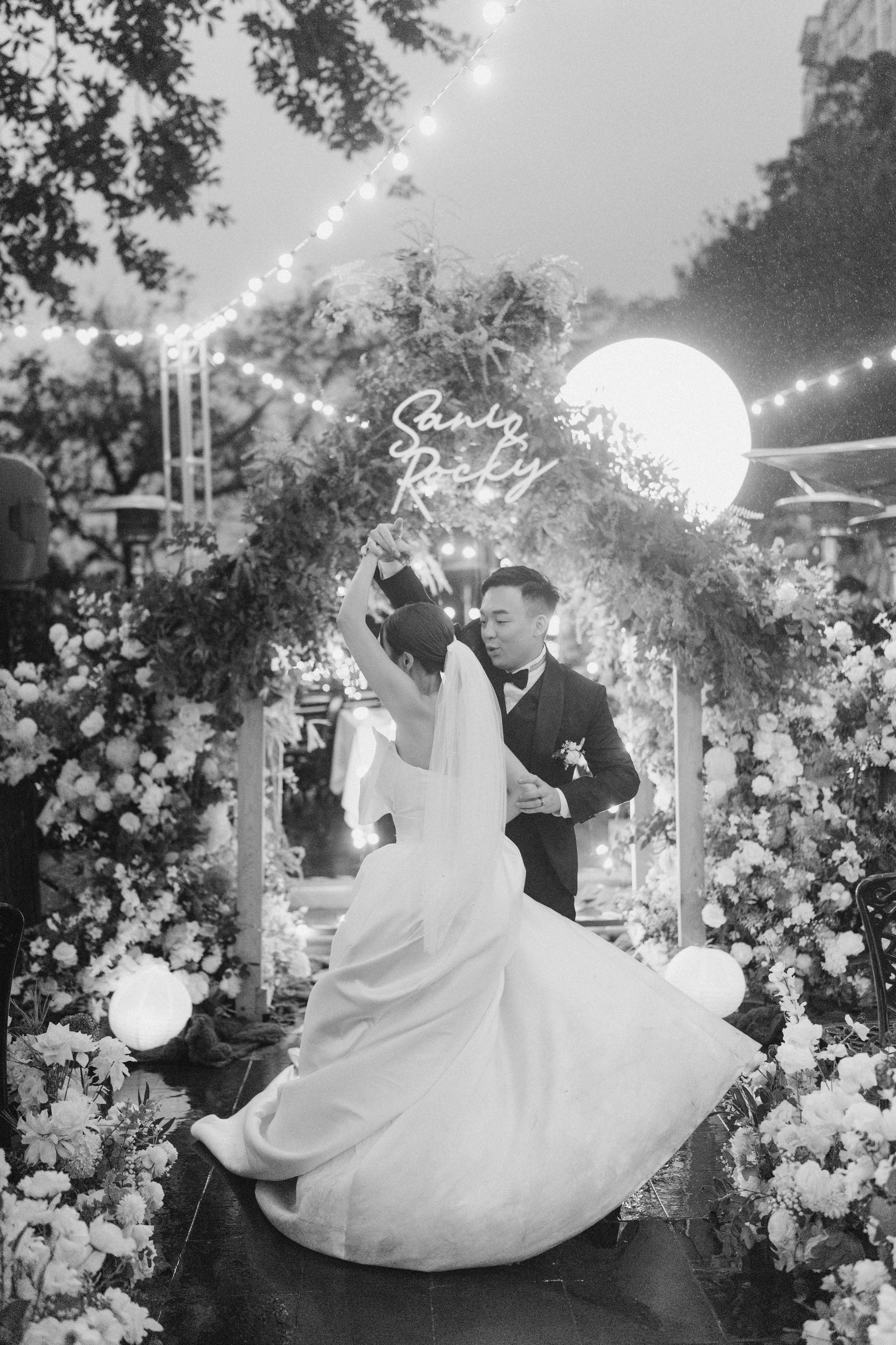 Bride and groom dance joyfully under a floral arch and string lights in a black and white wedding photo.