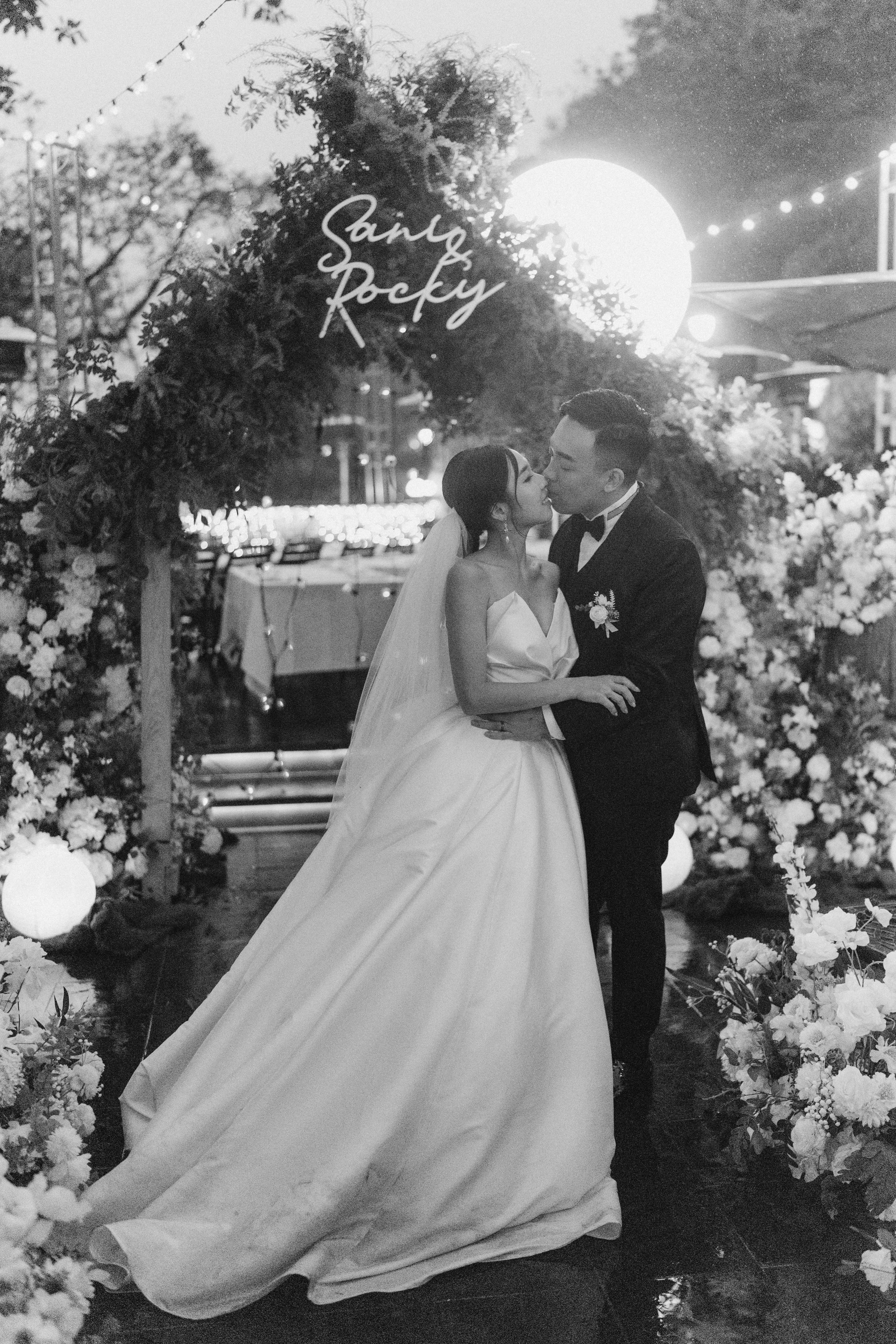 Bride and groom share a tender kiss under a neon sign and floral arch in a black and white wedding photo.