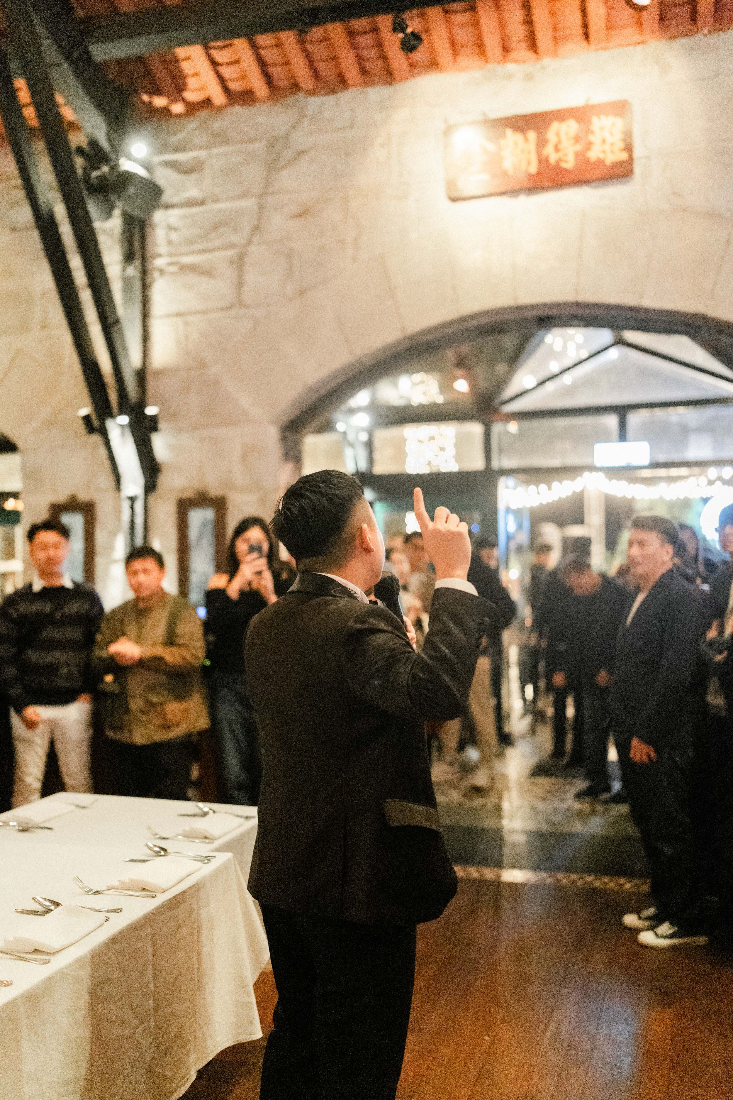 Groom addresses wedding guests inside a rustic stone venue decorated with warm ambient lighting.