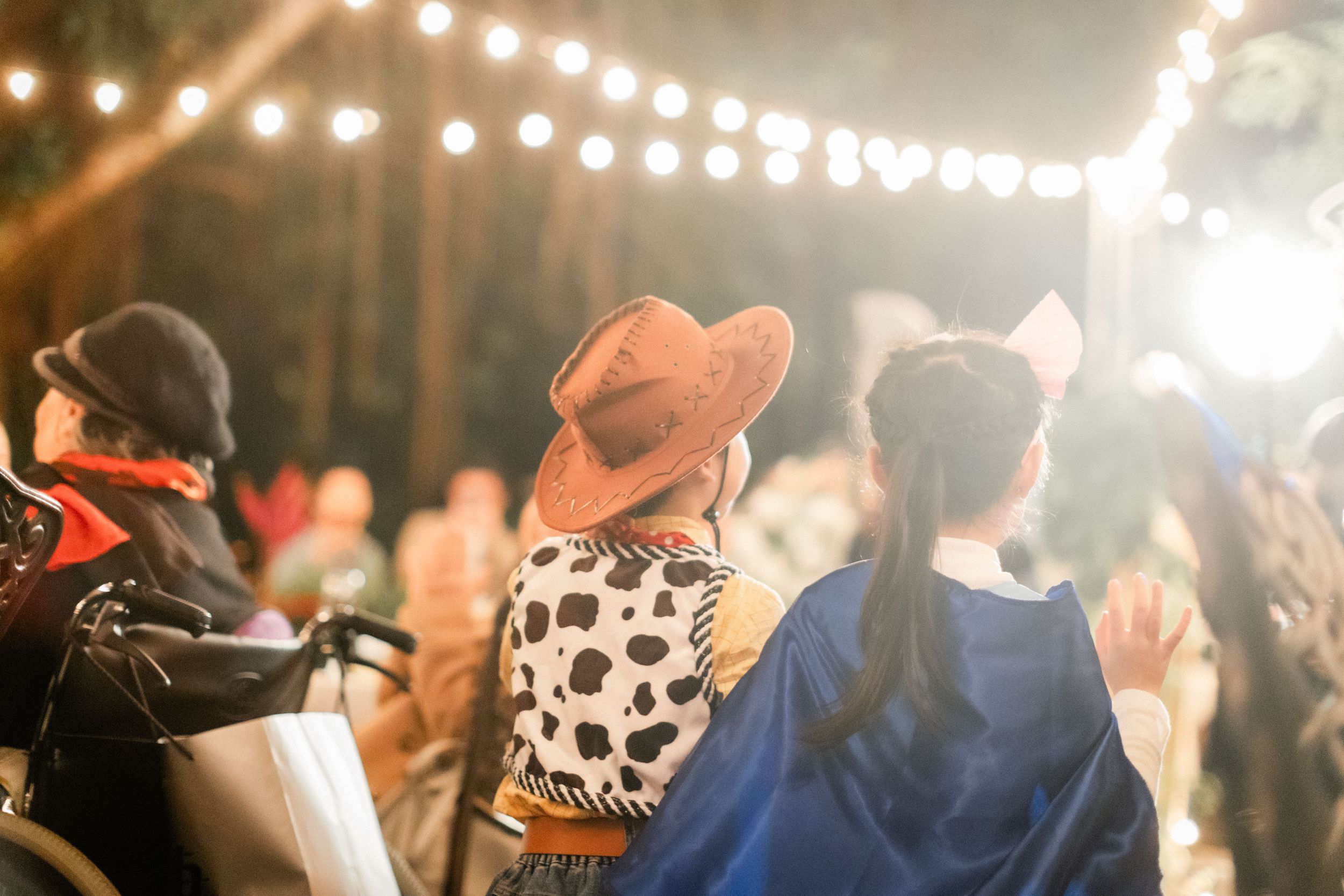 Wedding guests in colorful costumes enjoy an outdoor celebration under warm string lights at night.