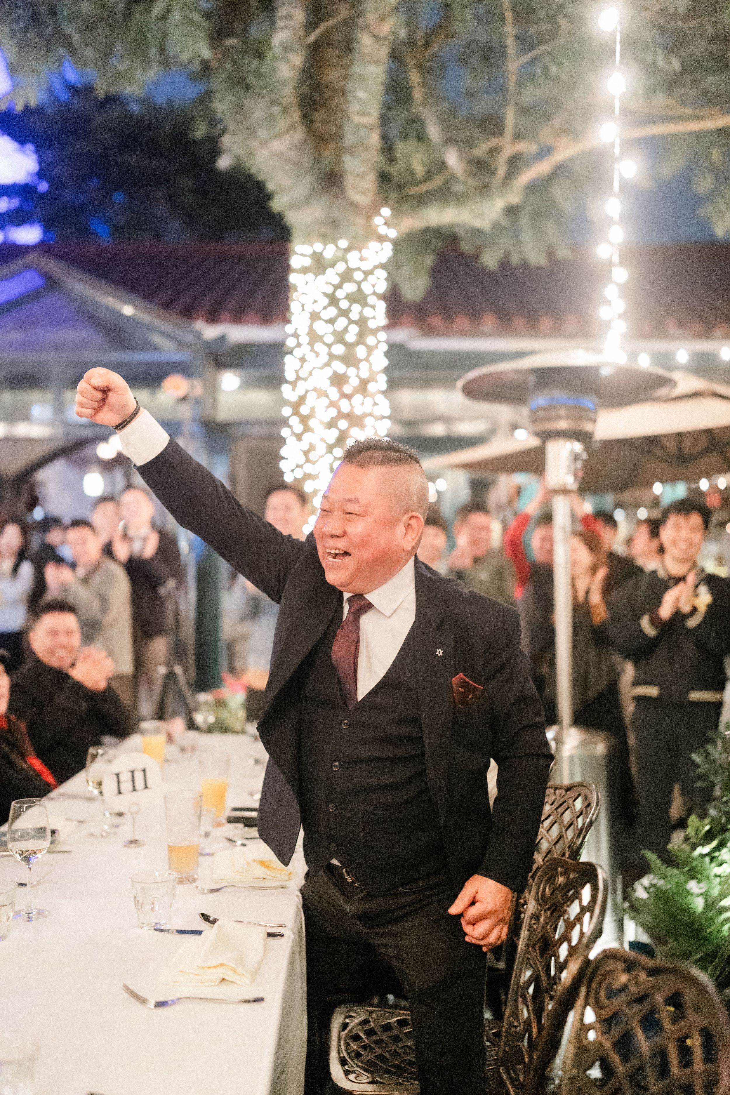 Excited man raises his fist in celebration at an outdoor wedding reception under twinkling lights.