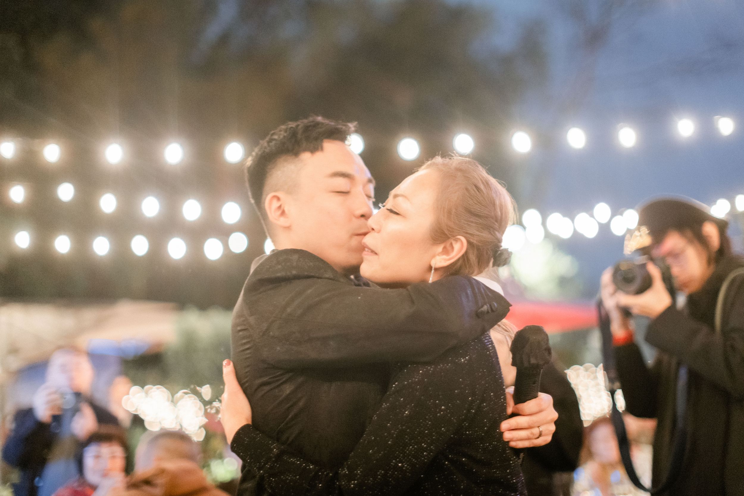 Couple shares a warm embrace and kiss on the dance floor under glowing string lights at a wedding.